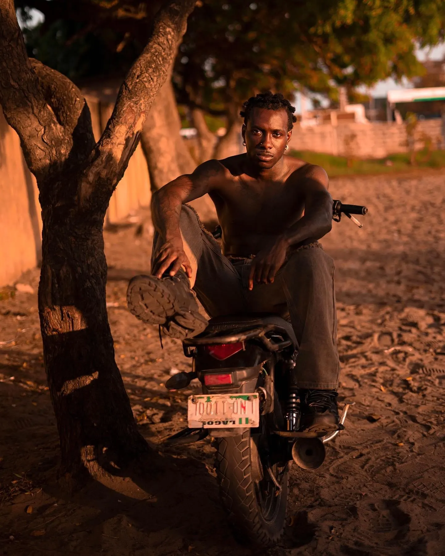 Seated on a Lagos-plated motorcycle in sand at dusk, one boot propped on the seat, gazing ahead.