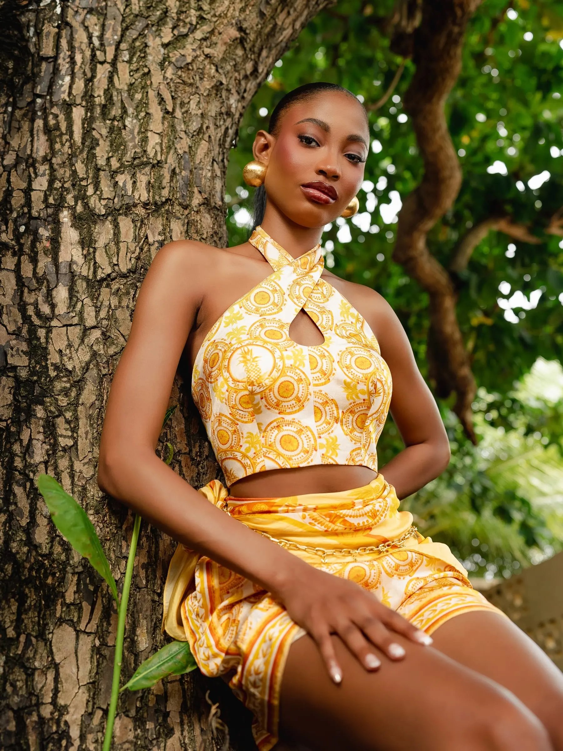 Model in a yellow cross-front halter crop top and matching printed skirt leans against the rough bark of a tree, gold hoop earrings and a direct gaze.