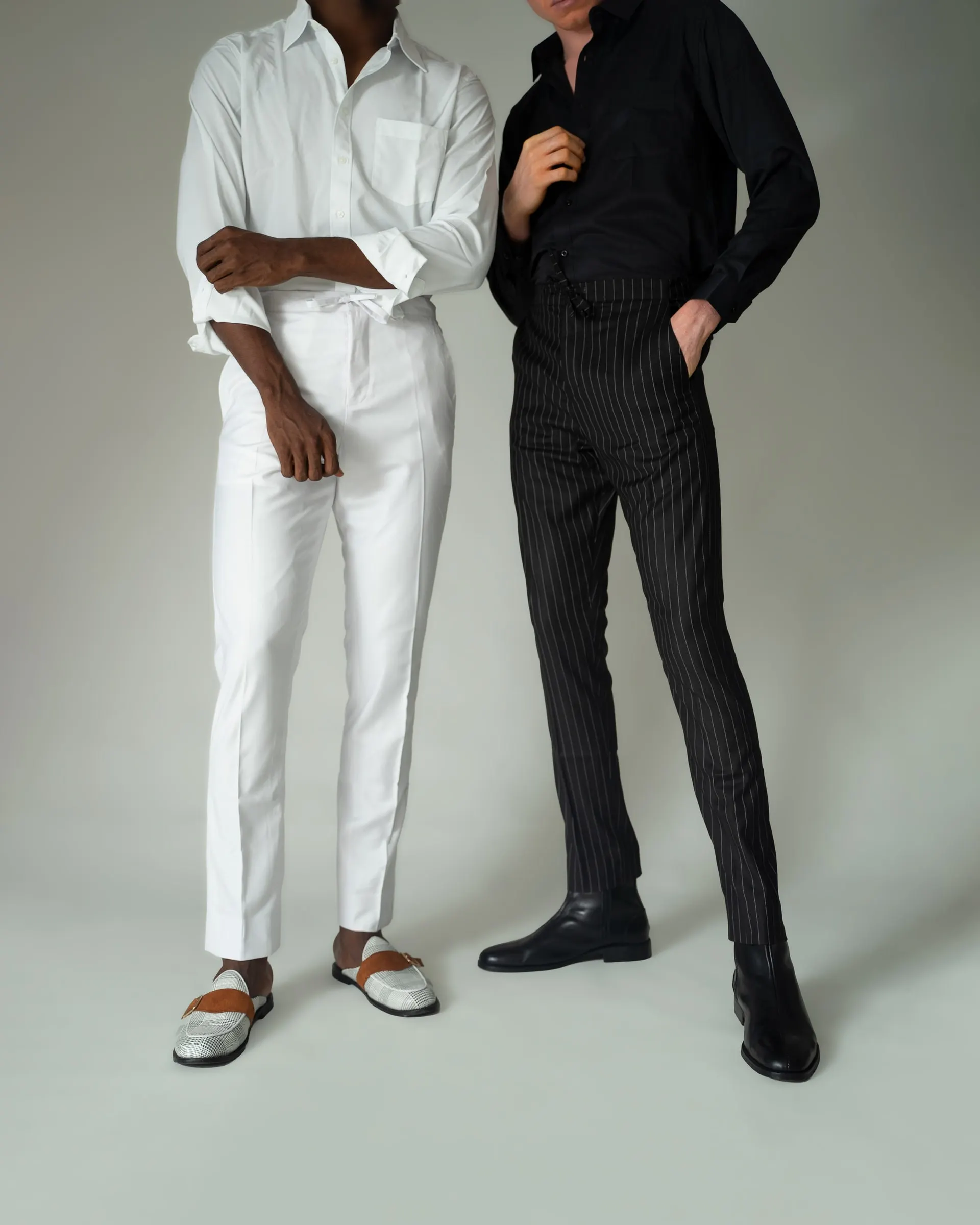 Two models from the shoulders down, one in a white shirt and trousers with cream loafers, the other in a black shirt and pinstripe trousers with black boots.