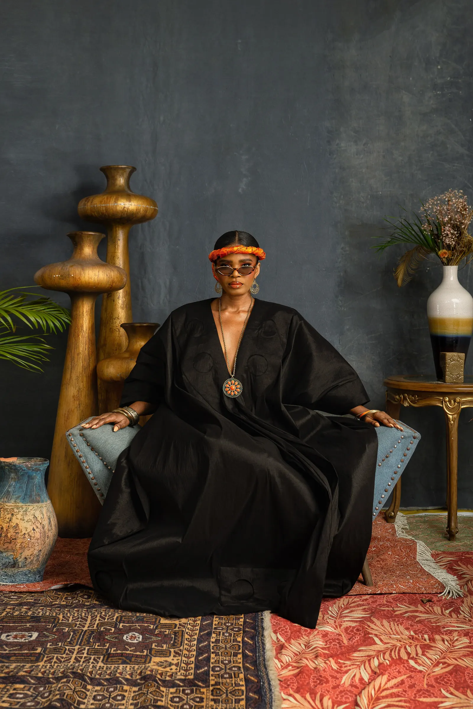 Model in a black boubou sits in a pale studded armchair between tall gold vessels, arms on the armrests, wearing an orange braided headband and tinted sunglasses, rugs layered on the floor.