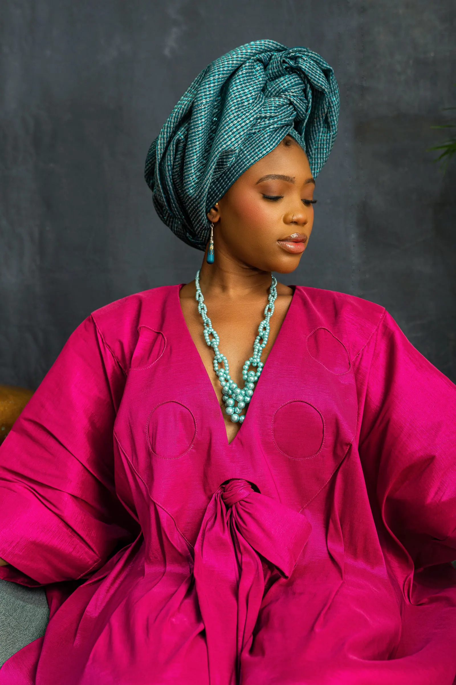 Close portrait of a model in a magenta-pink boubou with a teal patterned headwrap and a turquoise beaded necklace, eyes lowered, hands at the tie at the waist.