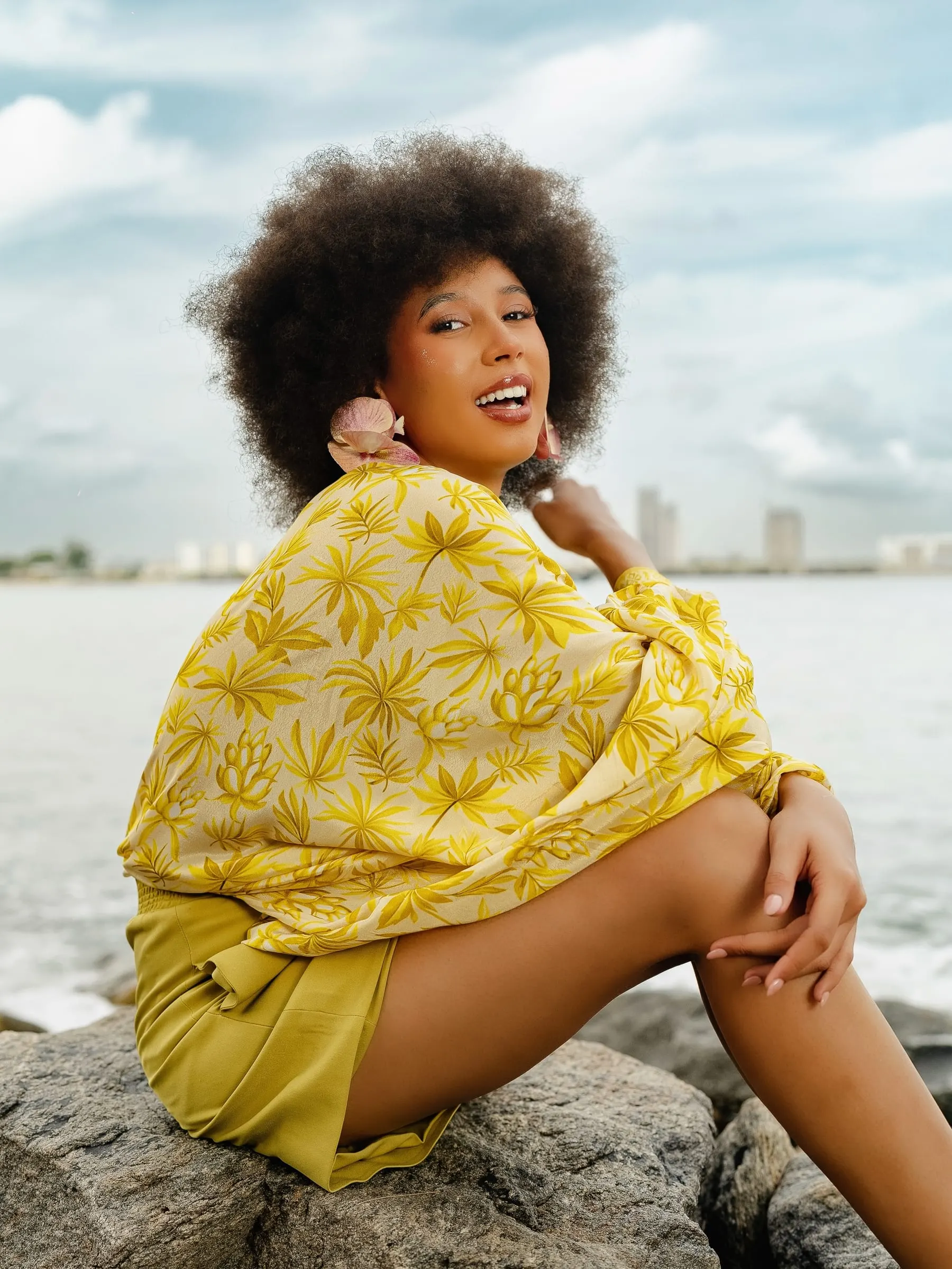 Model with a full afro laughs over her shoulder in a yellow palm-print shirt and olive shorts, seated on rocks with the sea and a city skyline across the water.