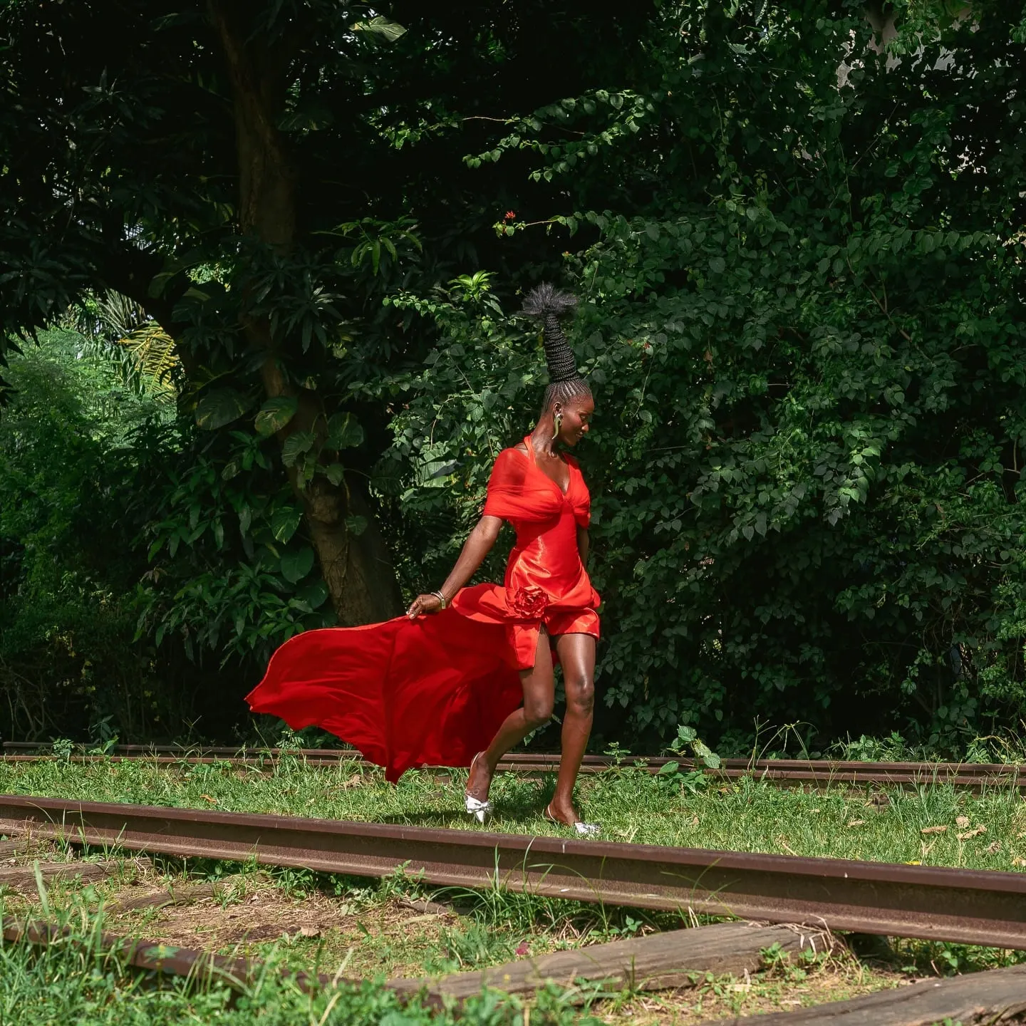 Model in a flowing red dress with a tall braided ponytail crosses disused rail tracks amid lush greenery.