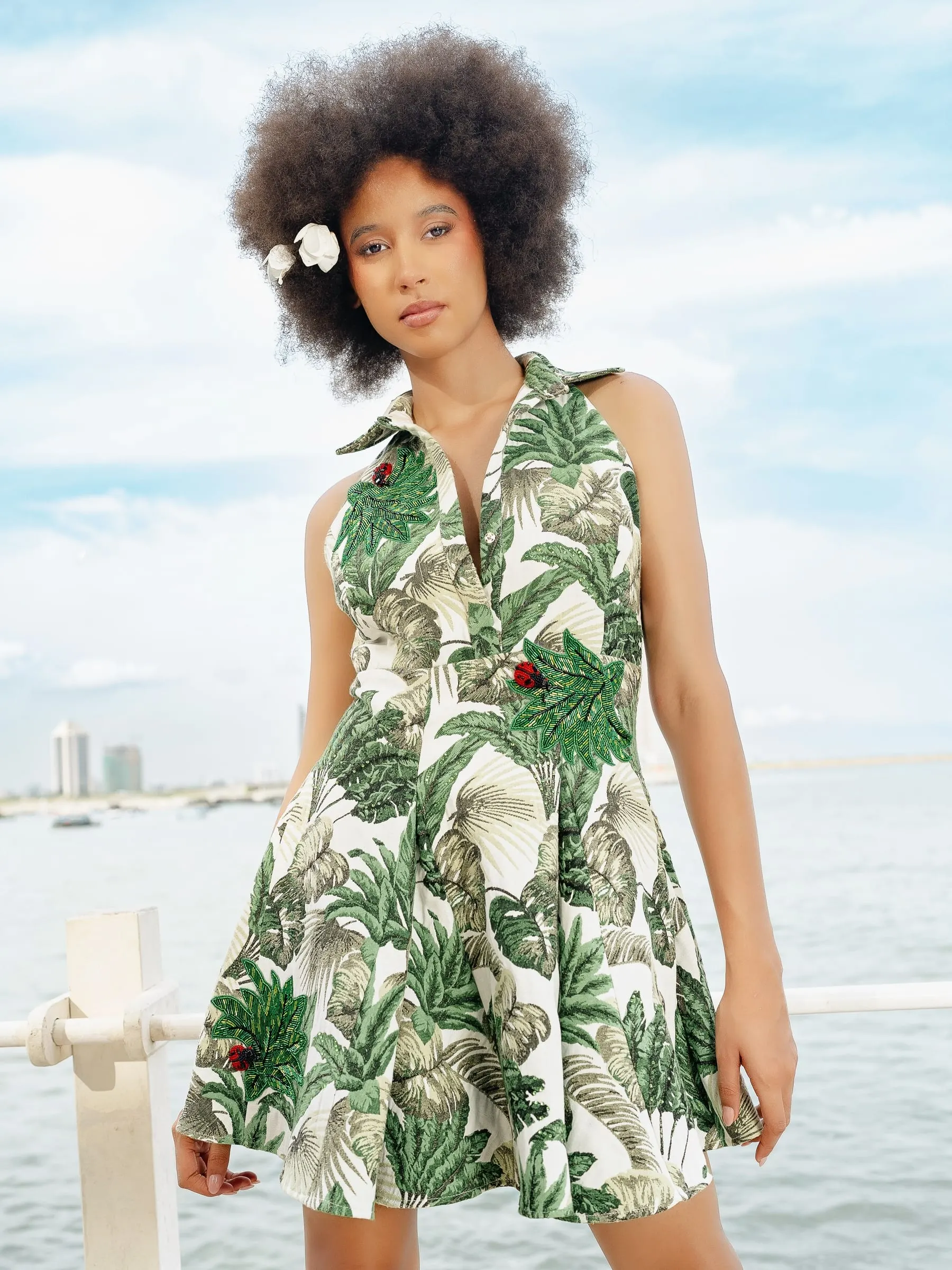 Model in a sleeveless green palm-print collared mini dress stands at a white railing by the water, a white flower in her afro and high-rises hazy across the bay.