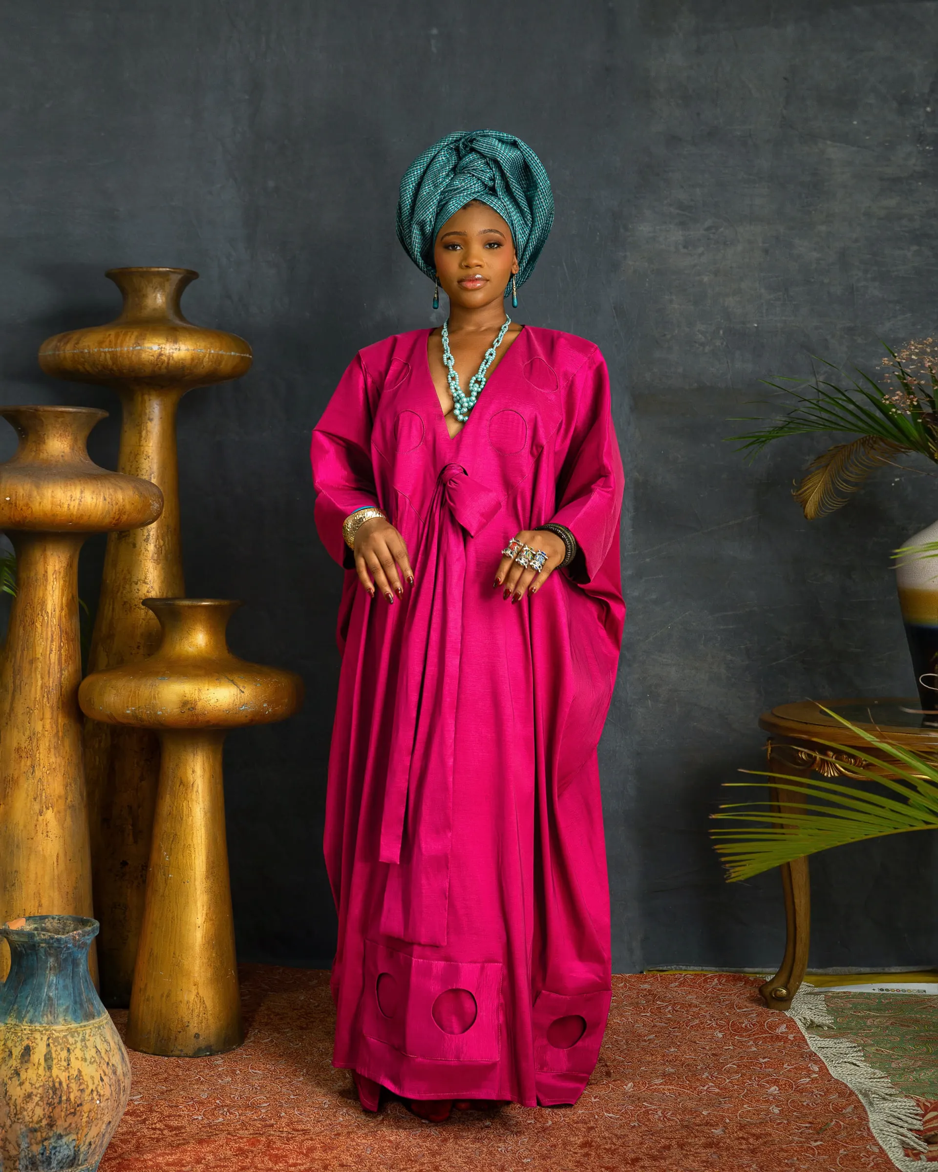 Model in a teal headwrap and turquoise necklace stands full length in a magenta-pink boubou with circular motifs at the hem, gold vessels to the left and palm fronds to the right.