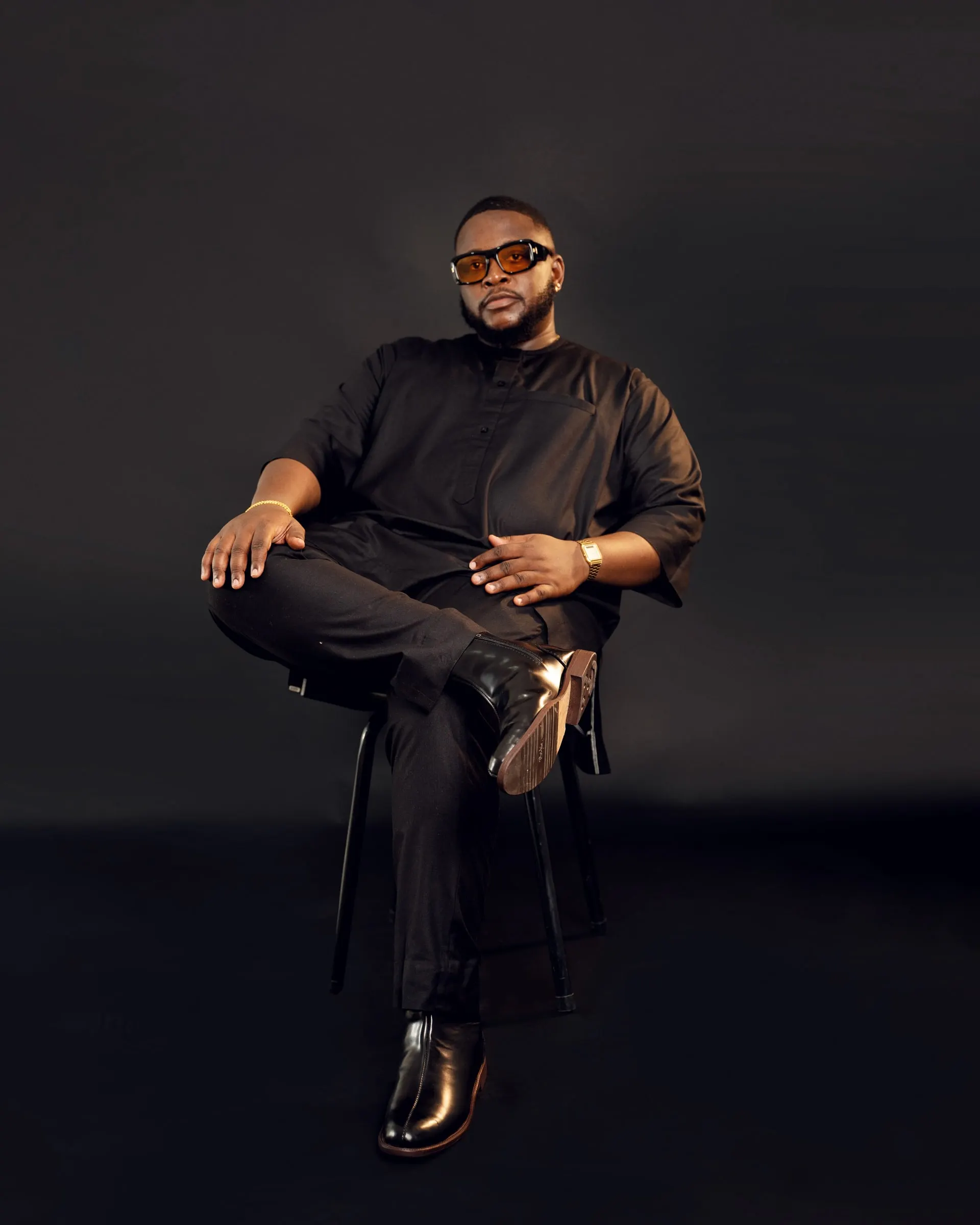 Man in a black tunic set and amber-tinted glasses sits on a stool with one leg crossed against a dark studio backdrop.