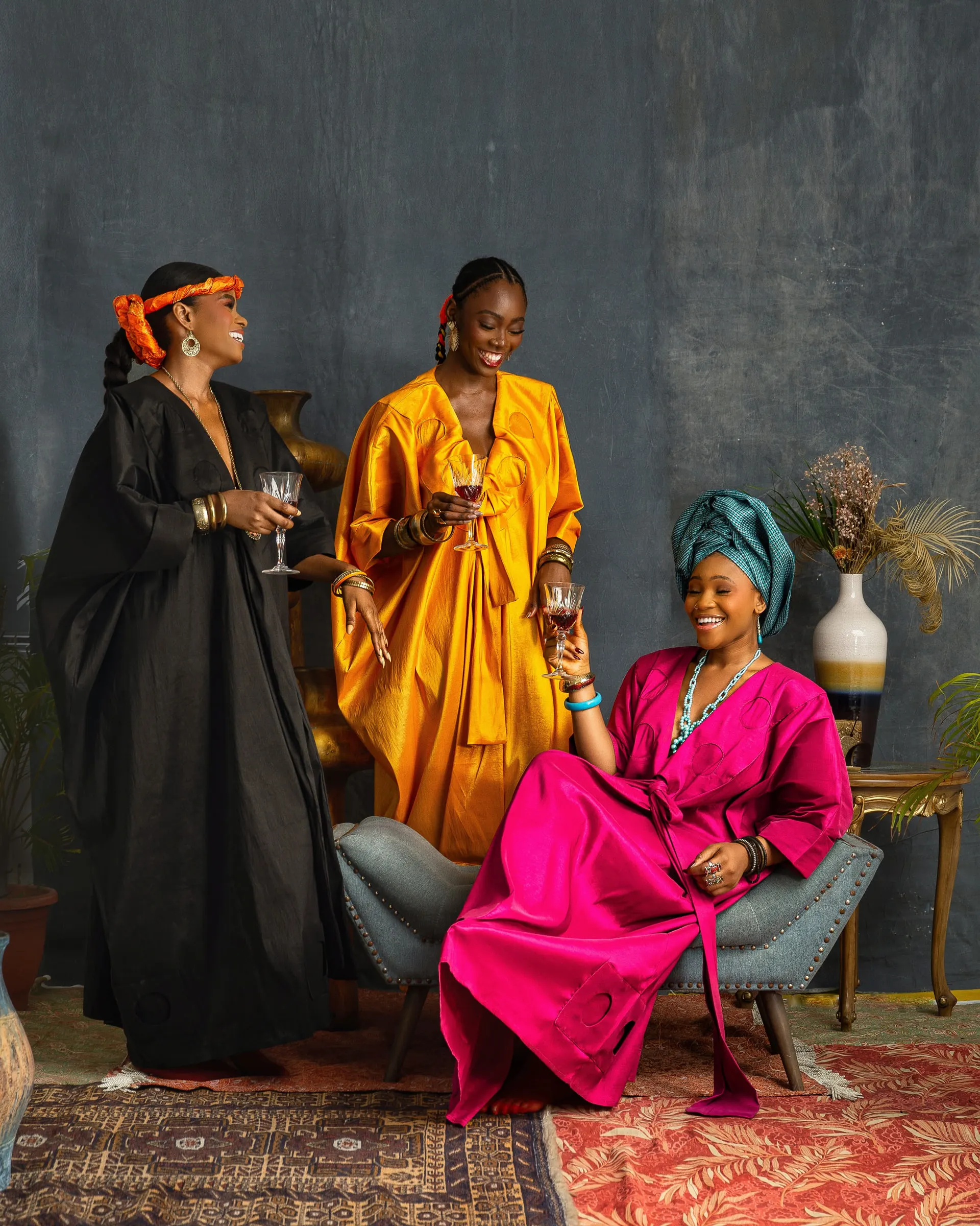 Three models with stemmed glasses: one in black laughs beside one in marigold orange, while a third in magenta pink with a teal headwrap sits in a pale armchair, a vase of dried grasses behind.