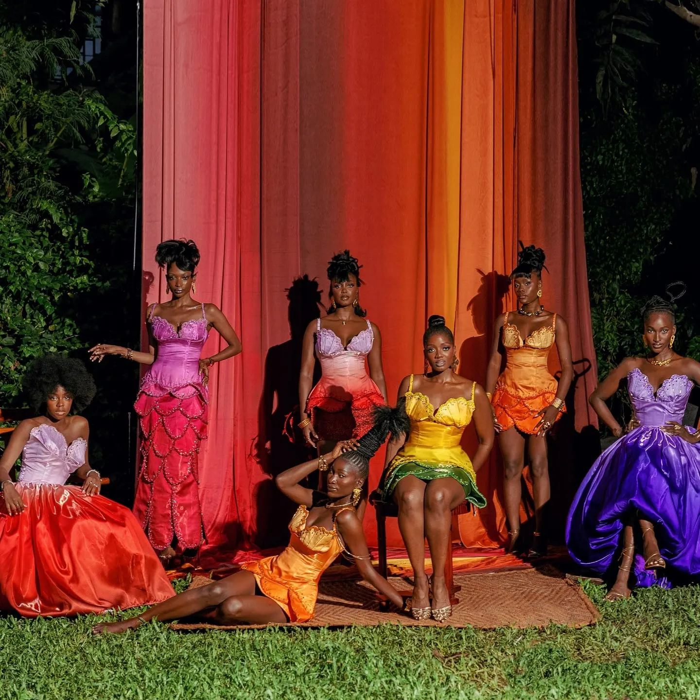 Seven models in gradient satin dresses gather on grass before a floor-to-ceiling sunset ombre backdrop at night.