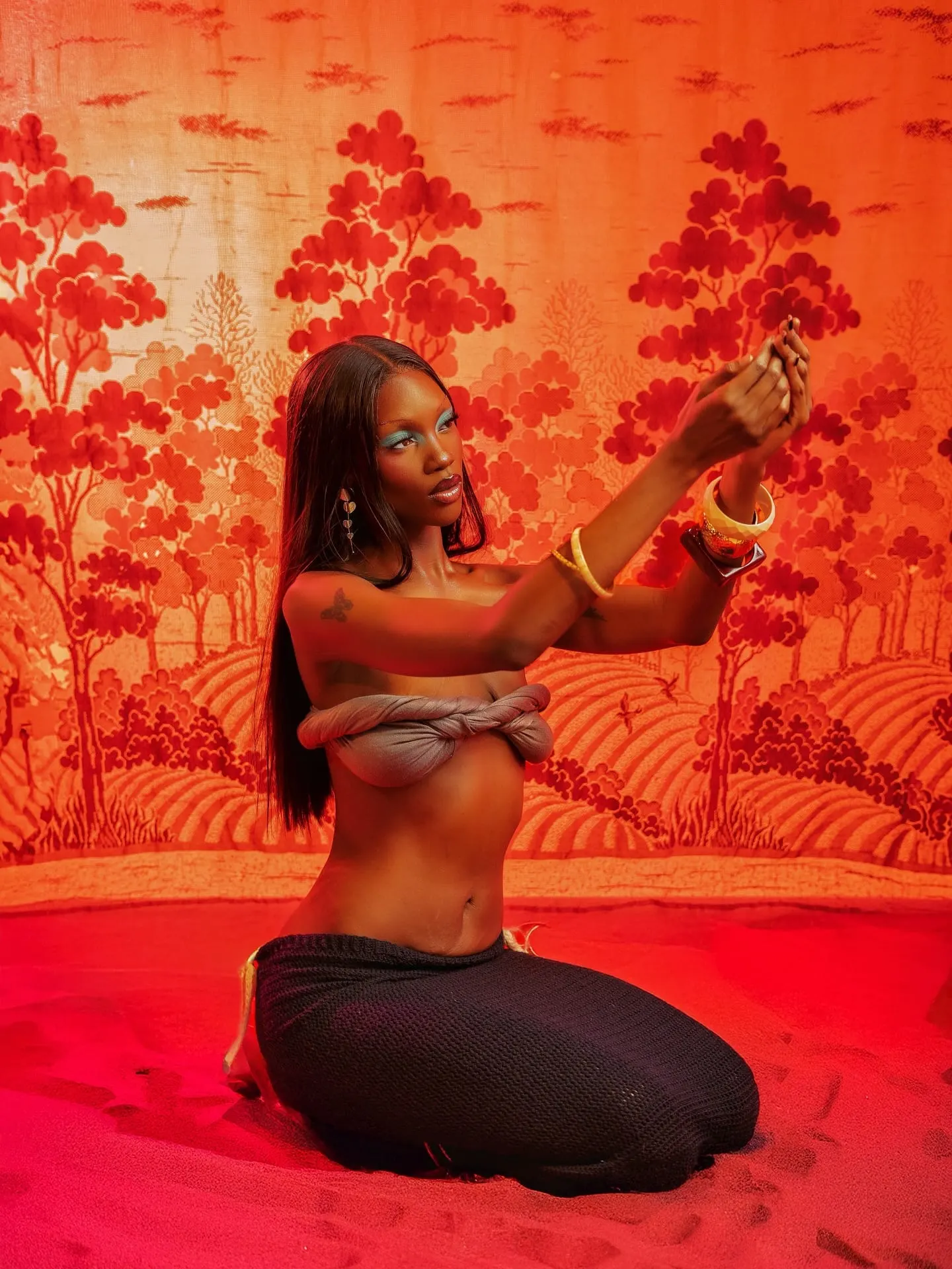 Kneeling model reaches both arms toward the light with cupped hands, gold bangles glinting against the red set.
