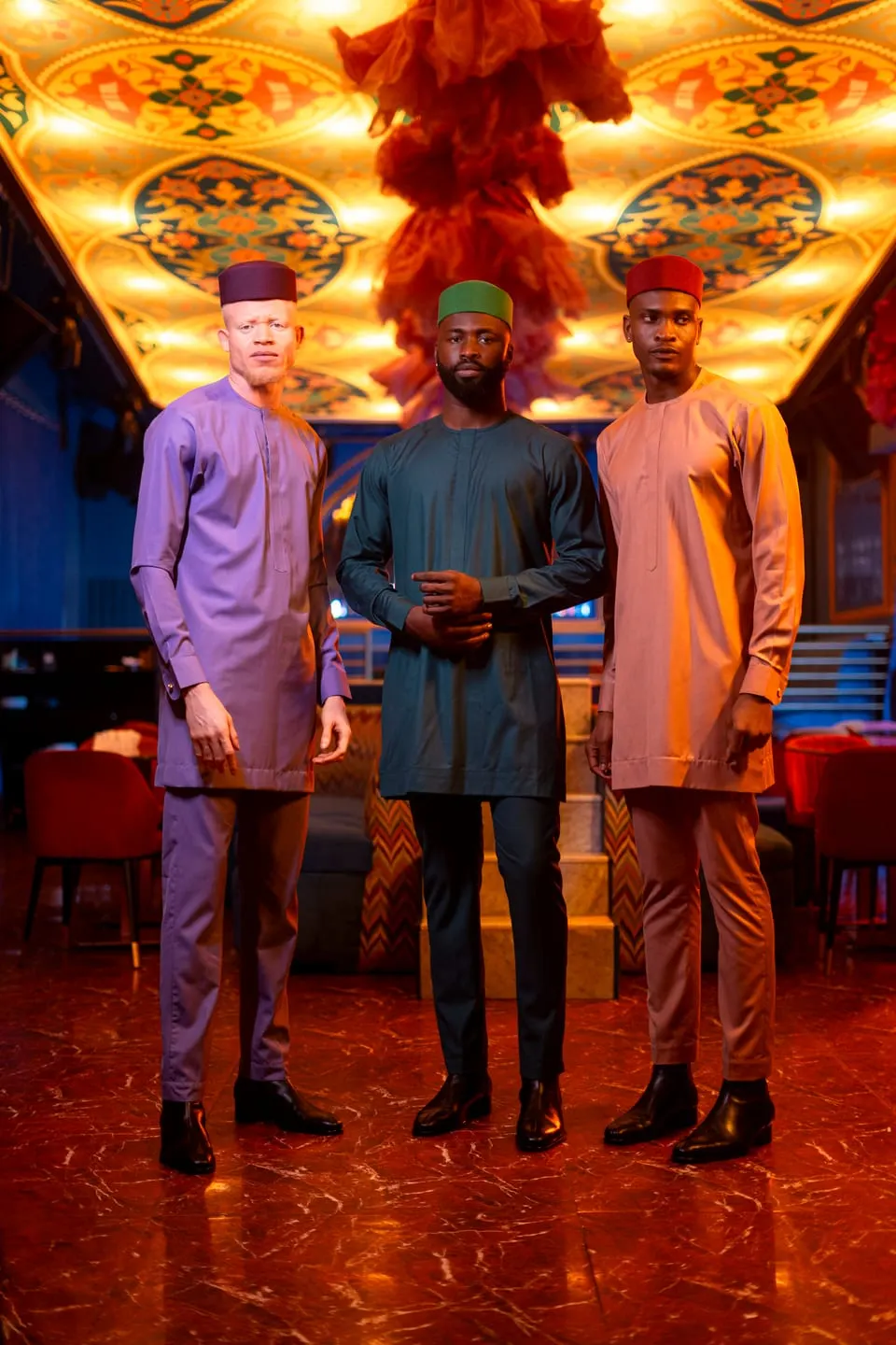 Three models in lilac, deep-green and blush tunic sets with matching caps stand in a lounge beneath an ornate gilded ceiling.