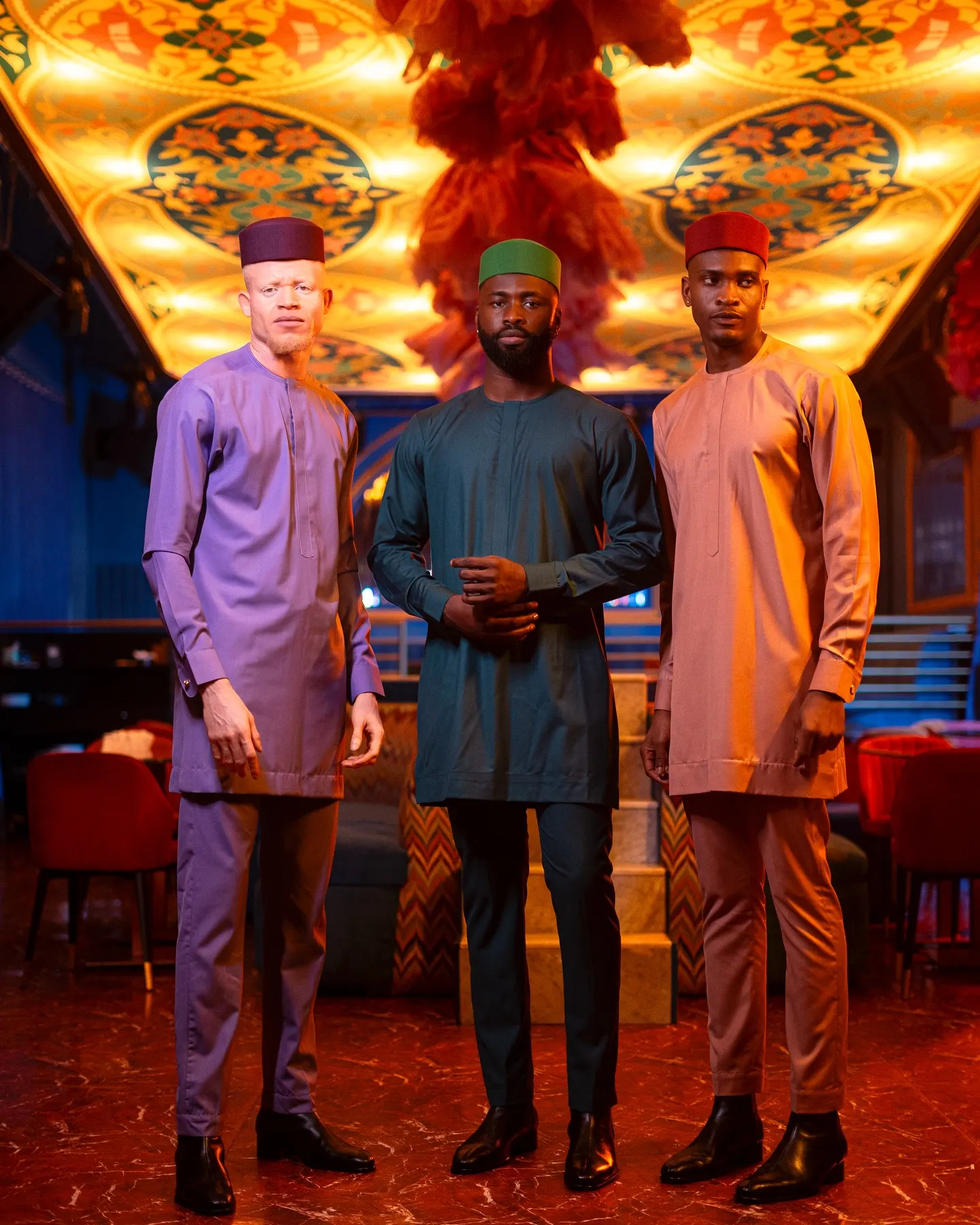 Three models in lilac, deep-green, and blush tunic sets with matching caps stand in a lounge beneath an ornate gilded ceiling.