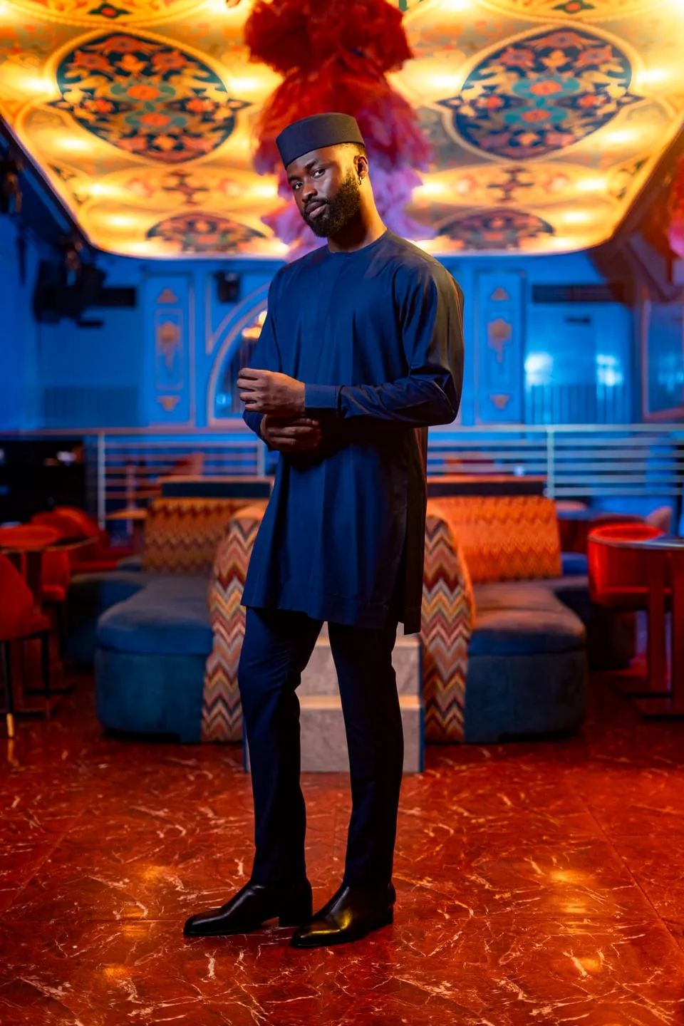 Model in a navy tunic and matching cap adjusts his cuff in a blue-lit lounge with red marble floors.