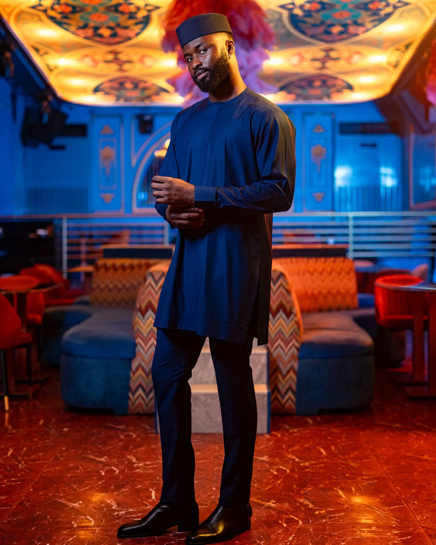 Model in a navy tunic and matching cap adjusts his cuff in a blue-lit lounge with red marble floors.