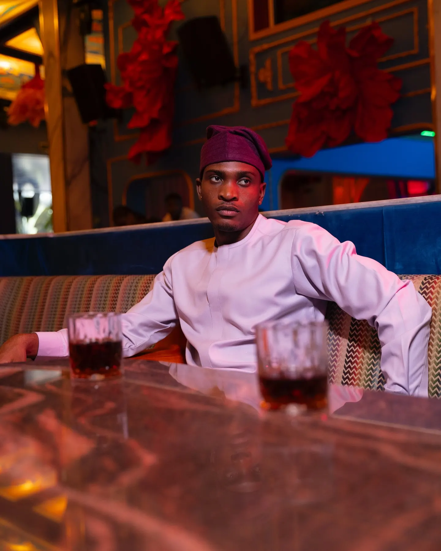 Model in a blush-pink tunic and maroon cap sits at a marble table, arm stretched along the booth, two glasses before him.