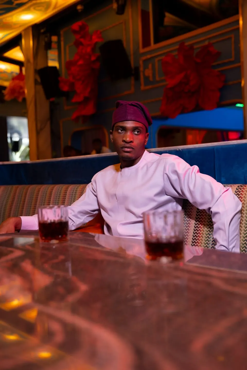 Model in a blush-pink tunic and maroon cap sits at a marble table, arm stretched along the booth, two glasses before him.