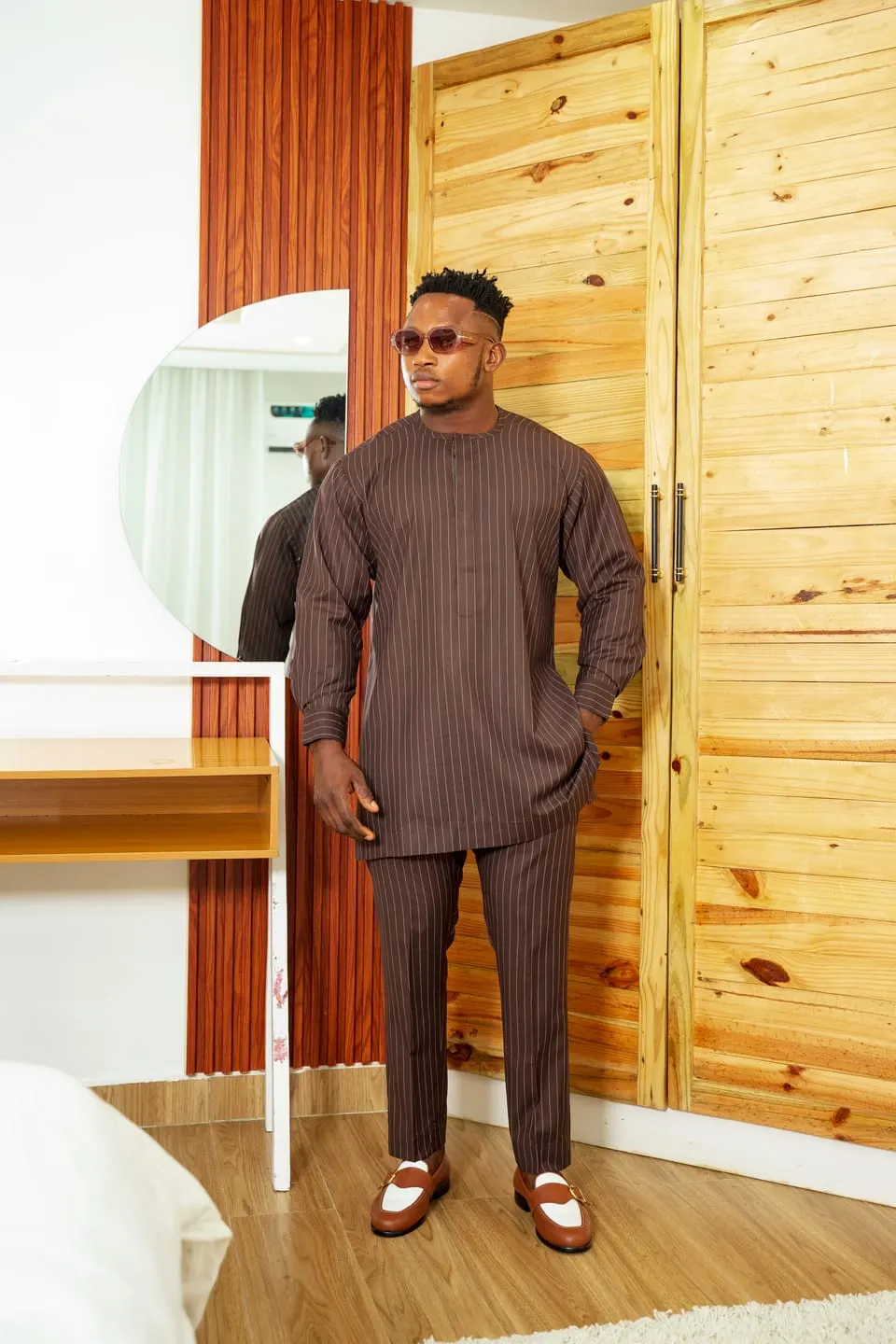 Model in a brown pinstripe tunic set and tinted glasses stands hand-in-pocket beside an arched mirror and pale wood wardrobe.