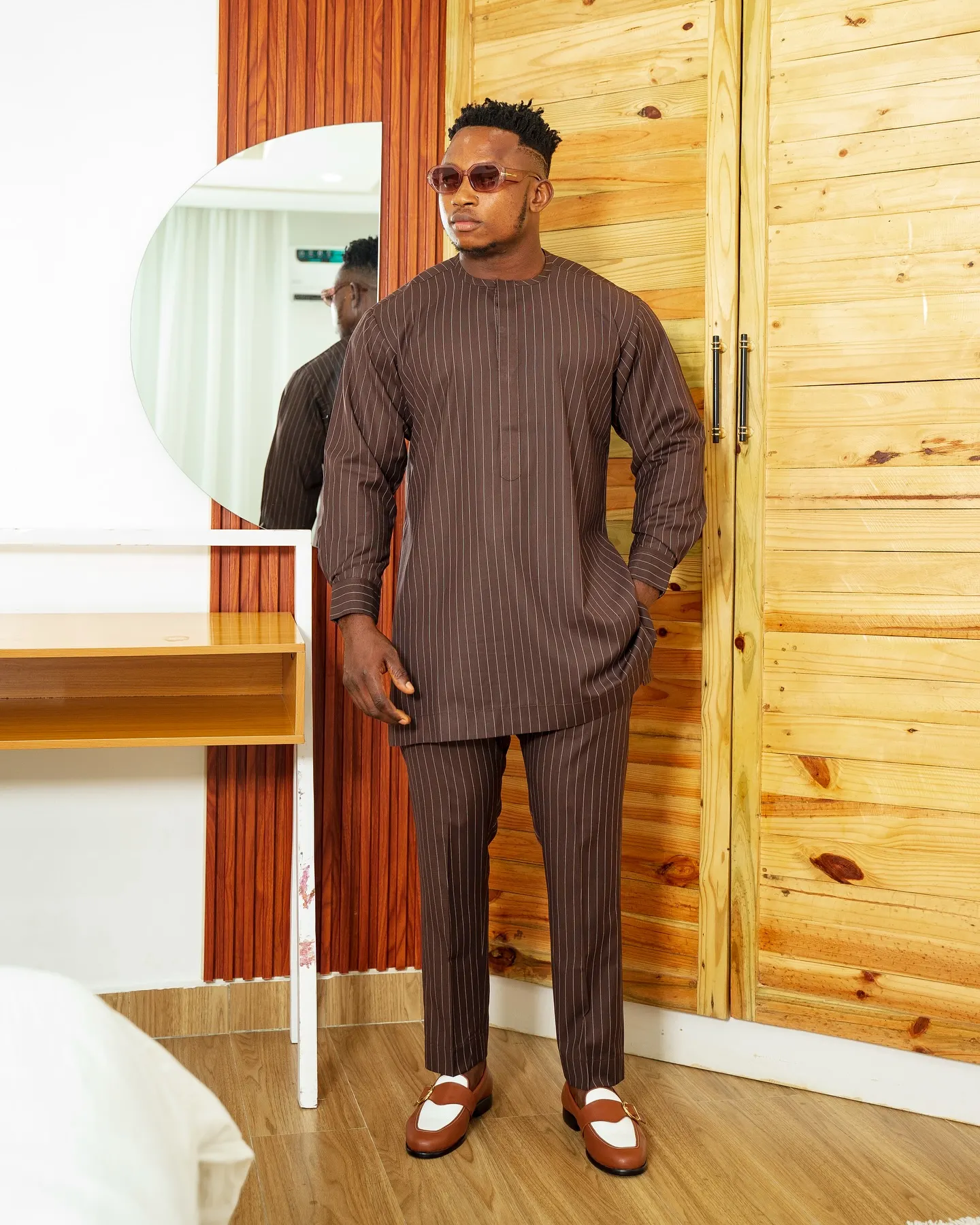 Model in a brown pinstripe tunic set and tinted glasses stands hand-in-pocket beside an arched mirror and pale wood wardrobe.