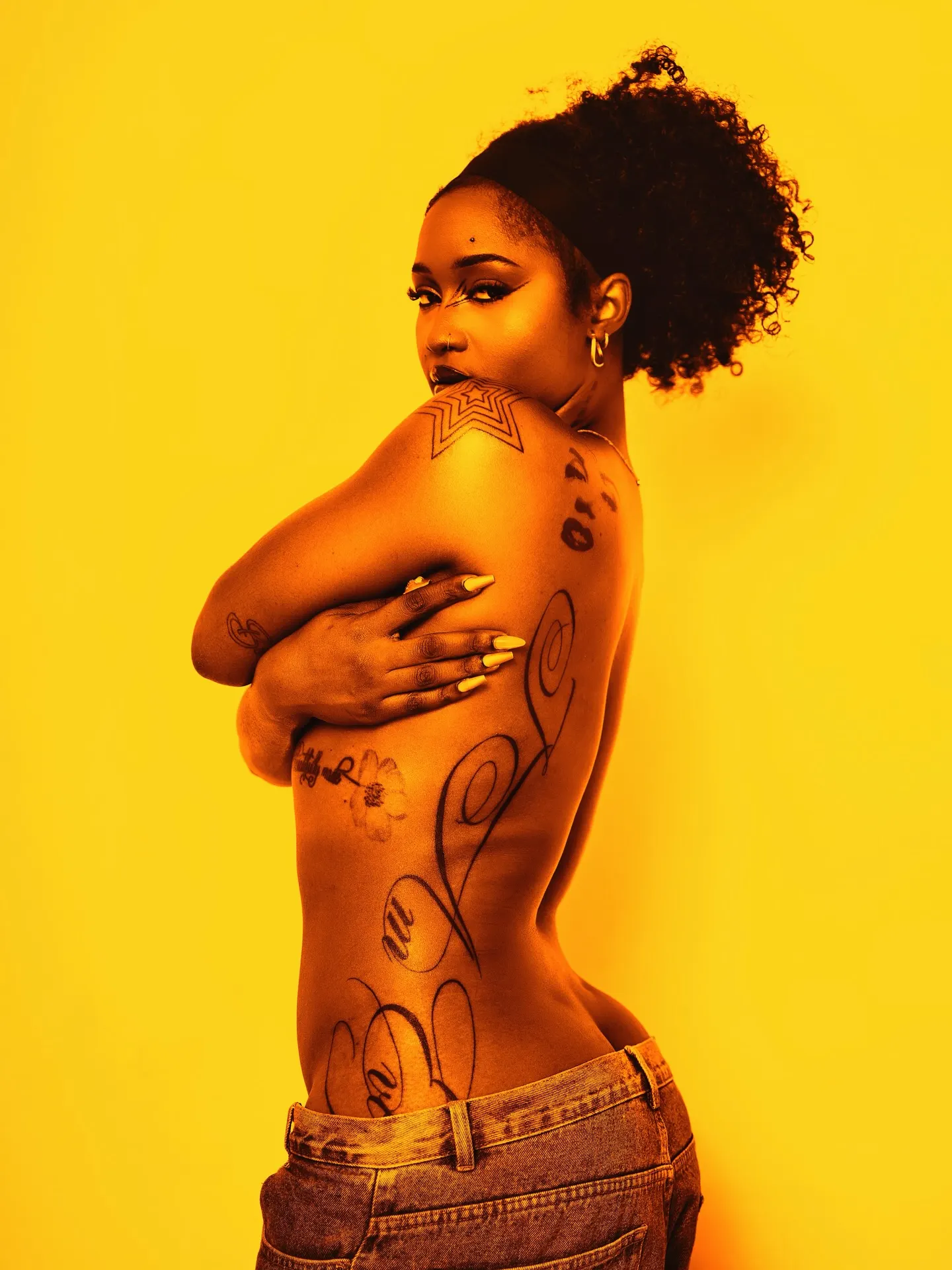 Tattooed model glancing over her shoulder with arms crossed over her bare back, jeans slung low, washed in monochrome yellow.