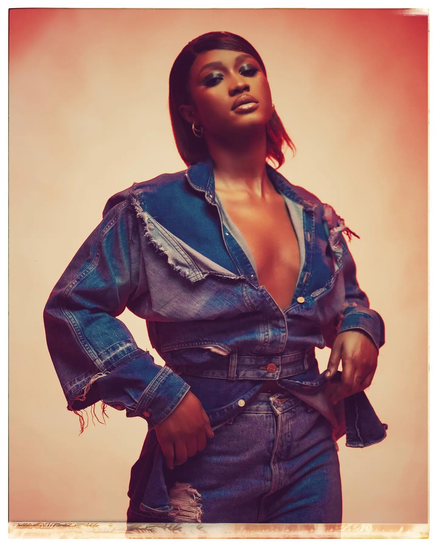 Model in a frayed patchwork denim jacket and jeans, hands on hips under warm red light, framed by a Polaroid border.