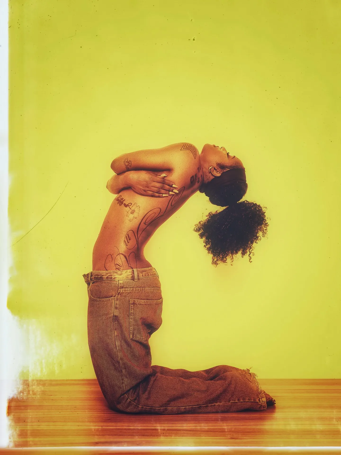 Kneeling model arching backward in baggy jeans, curls spilling toward the floor, her tattooed torso lit against a chartreuse wall.