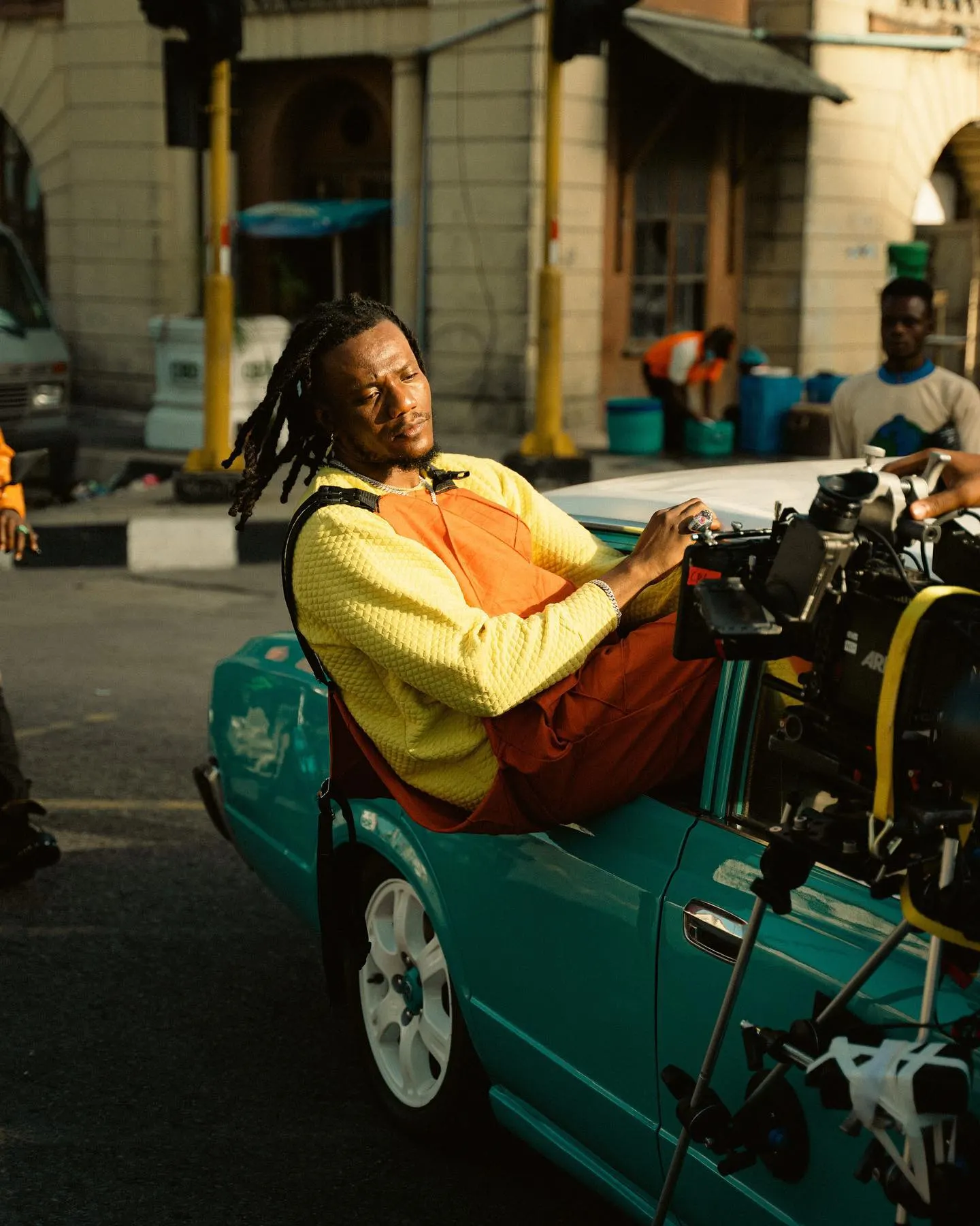 Pheelz in a yellow knit sweater and orange overalls reclining across the trunk of a teal convertible on a sunlit Lagos street, a rigged cinema camera in the foreground.