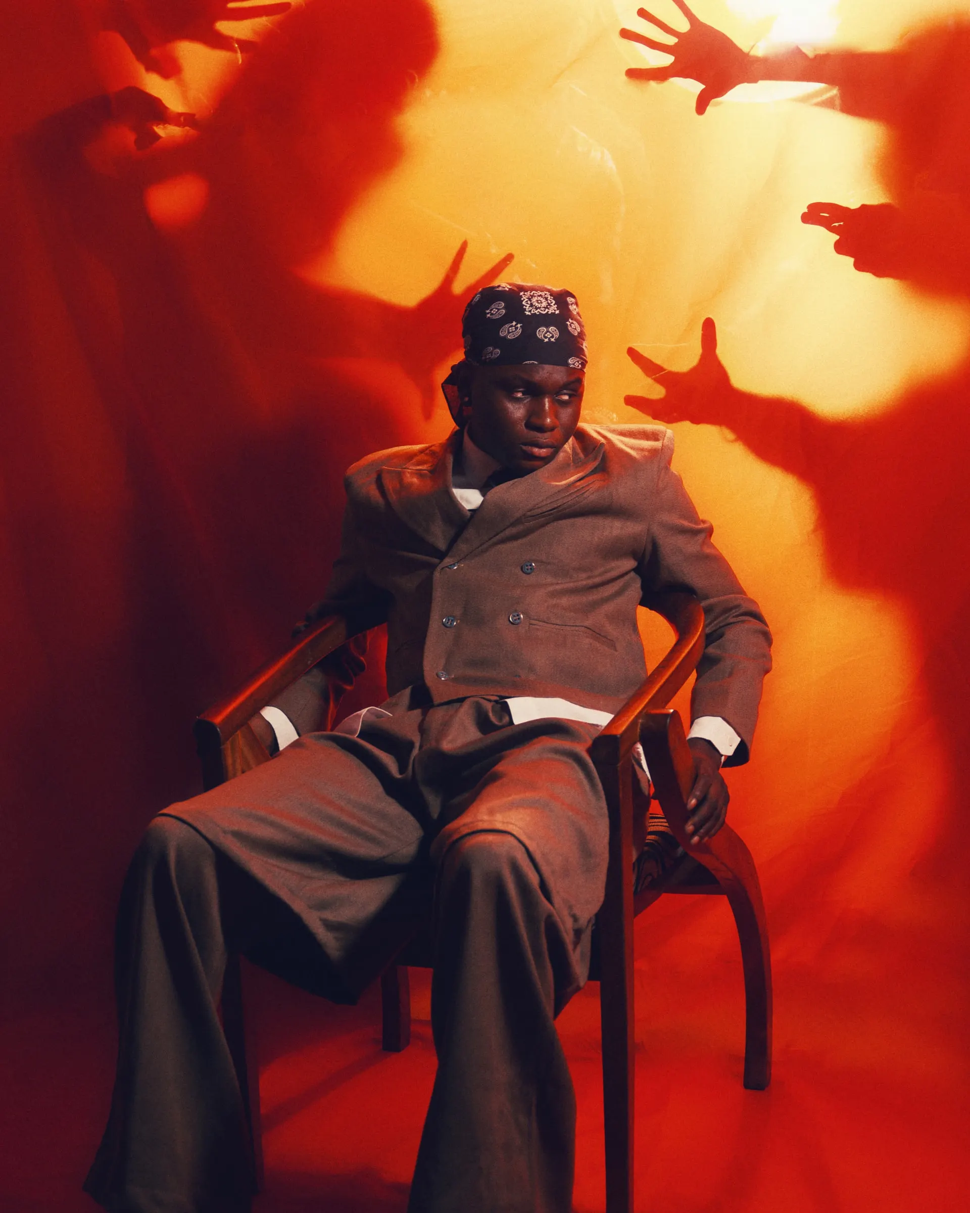 Musician in a grey double-breasted suit and paisley bandana slouched in a wooden chair, shadowy hands reaching toward him across a red-orange backdrop.