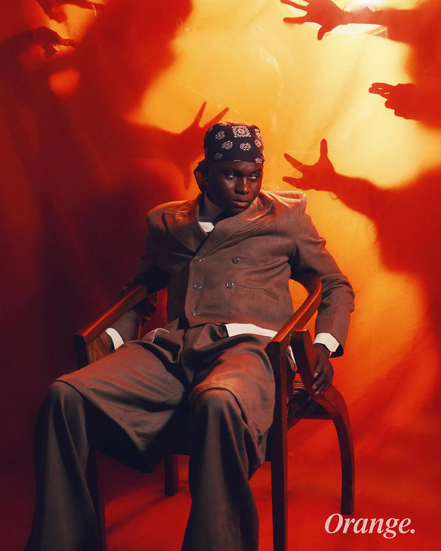 Musician in a grey double-breasted suit and paisley bandana slouched in a wooden chair, shadowy hands reaching toward him across a red-orange backdrop.