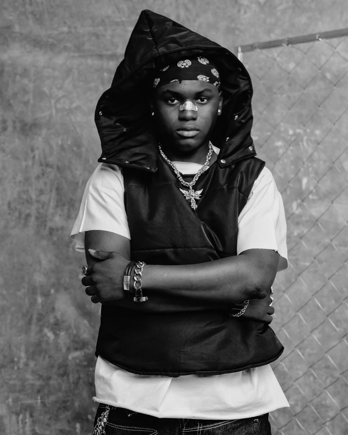 Black-and-white portrait of Ayo Maff with arms folded, hood up over his bandana, silver cross pendant resting on his puffer vest in front of the chain-link fence.