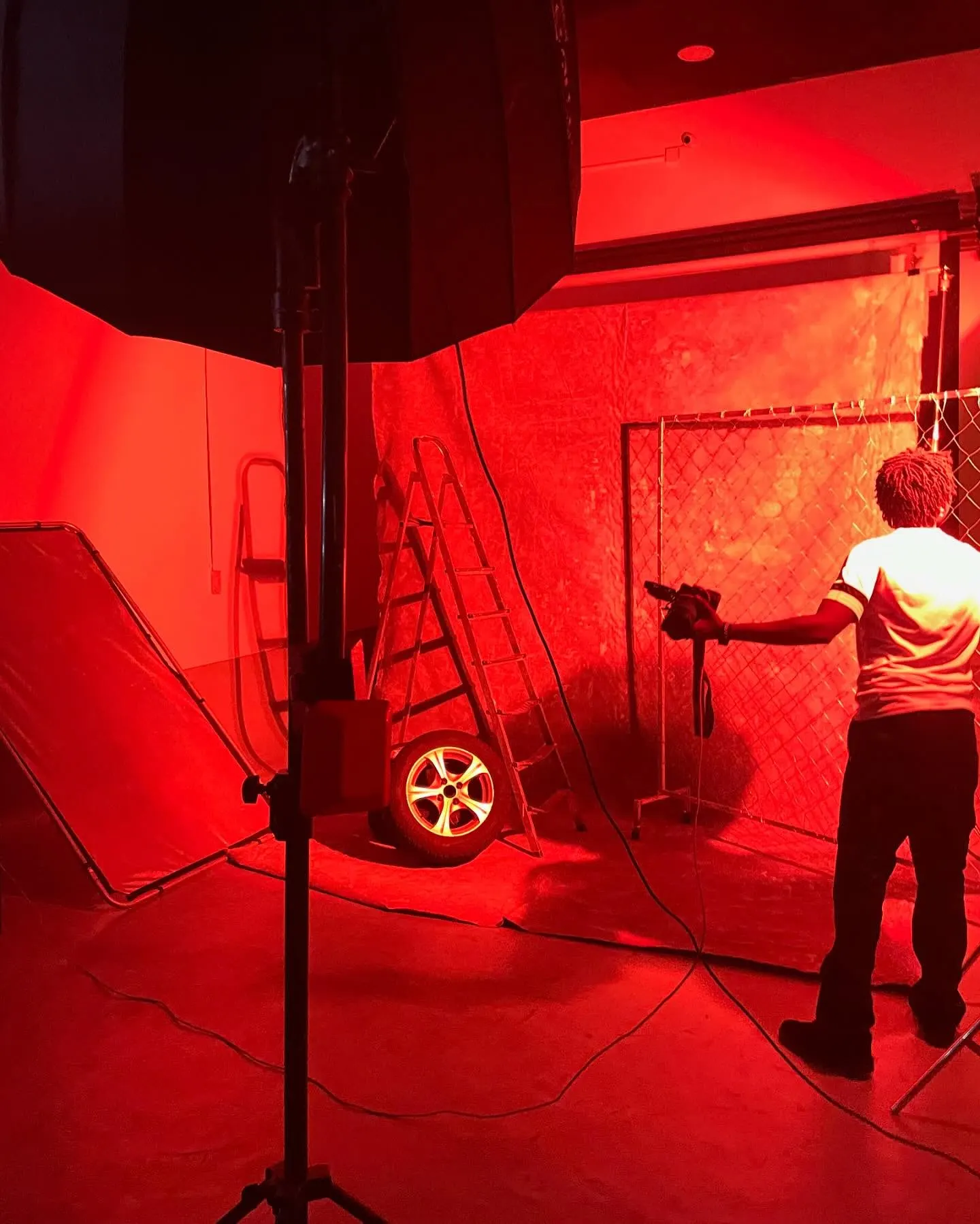 Behind the scenes bathed in red light — a crew member adjusts gear beside a ladder, lone tyre and chain-link fence on the studio set.