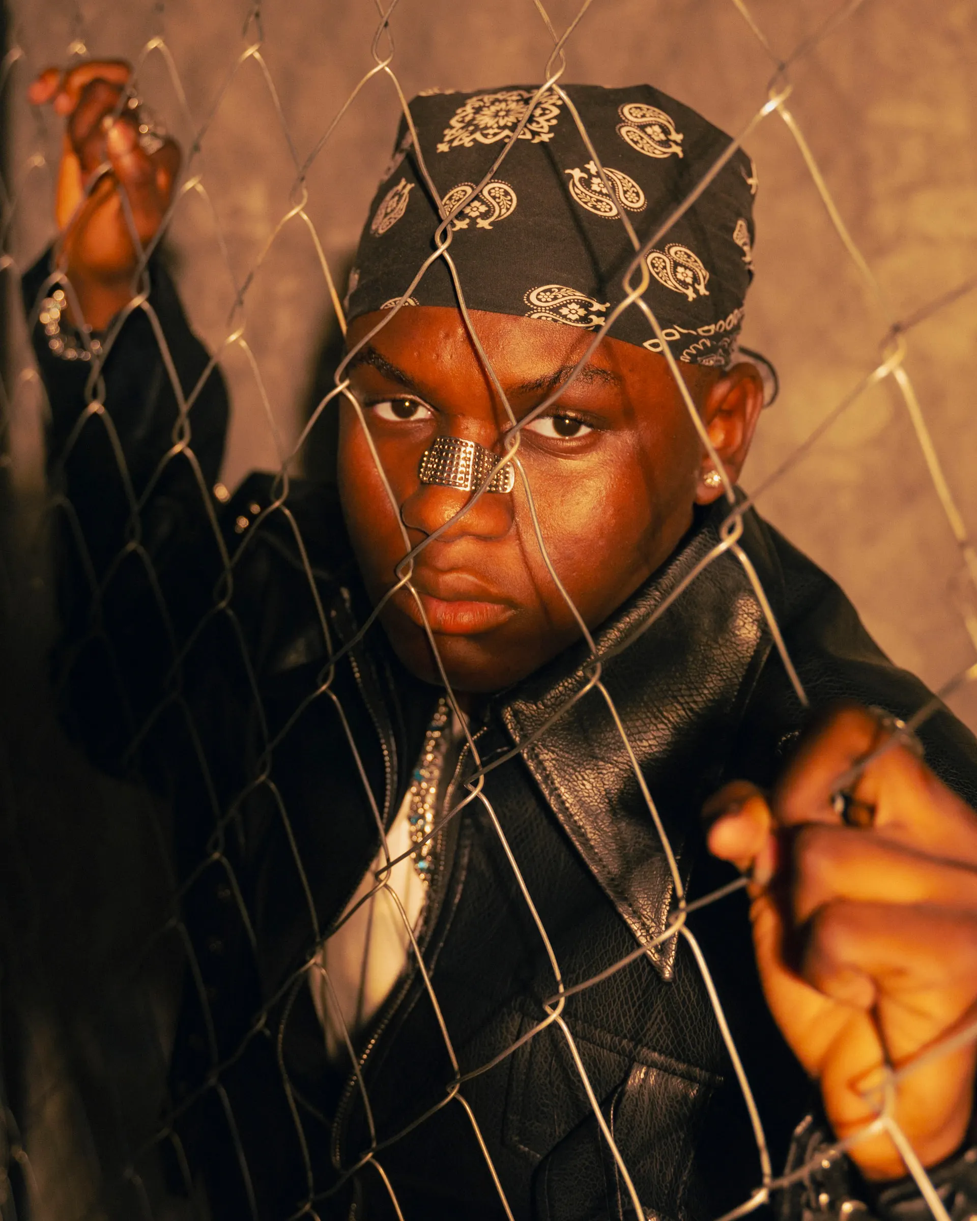 Close-up of a musician in a dark paisley bandana and leather jacket gripping a chain-link fence, face pressed toward the wire under warm amber light.
