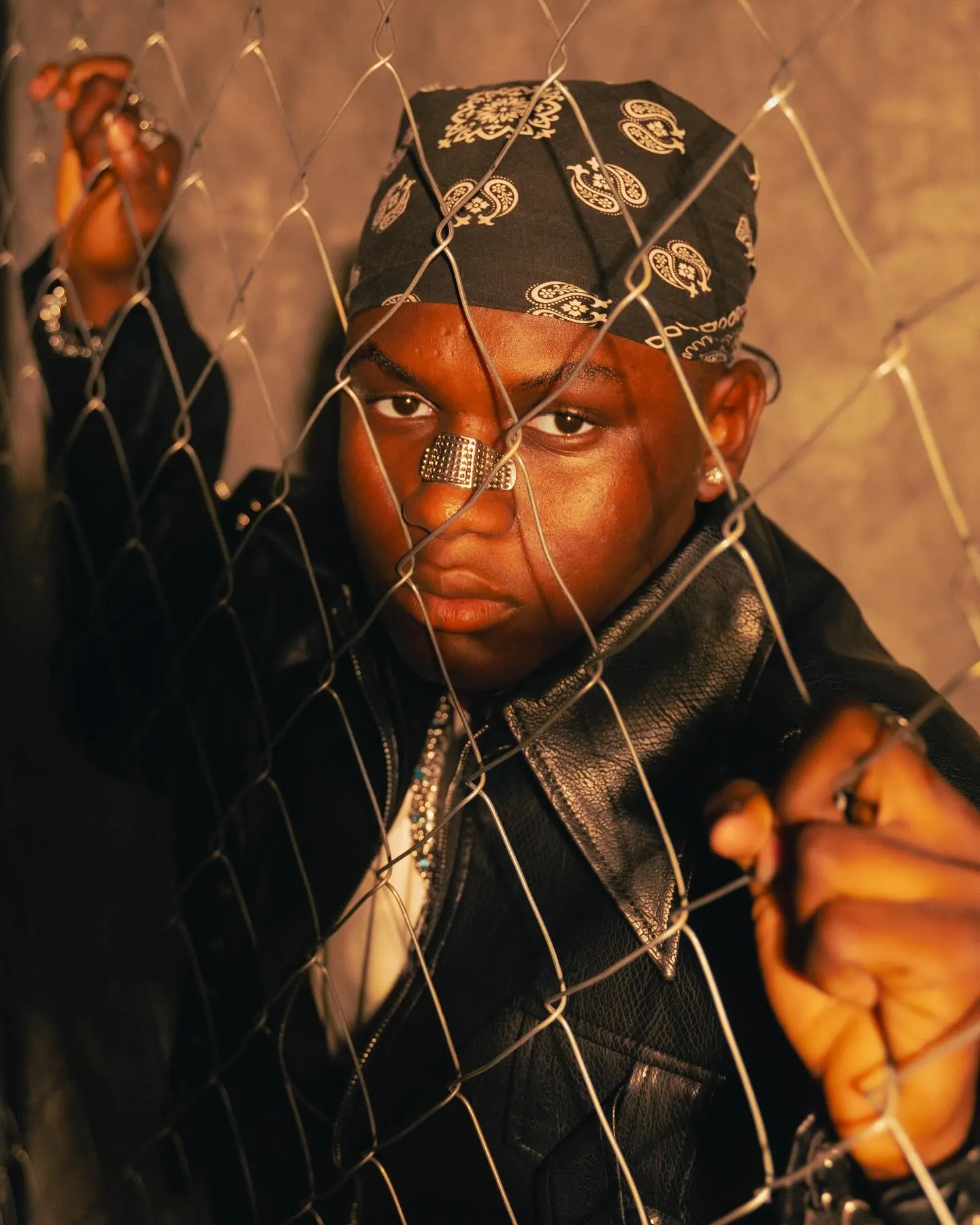 Close-up of a musician in a dark paisley bandana and leather jacket gripping a chain-link fence, face pressed toward the wire under warm amber light.
