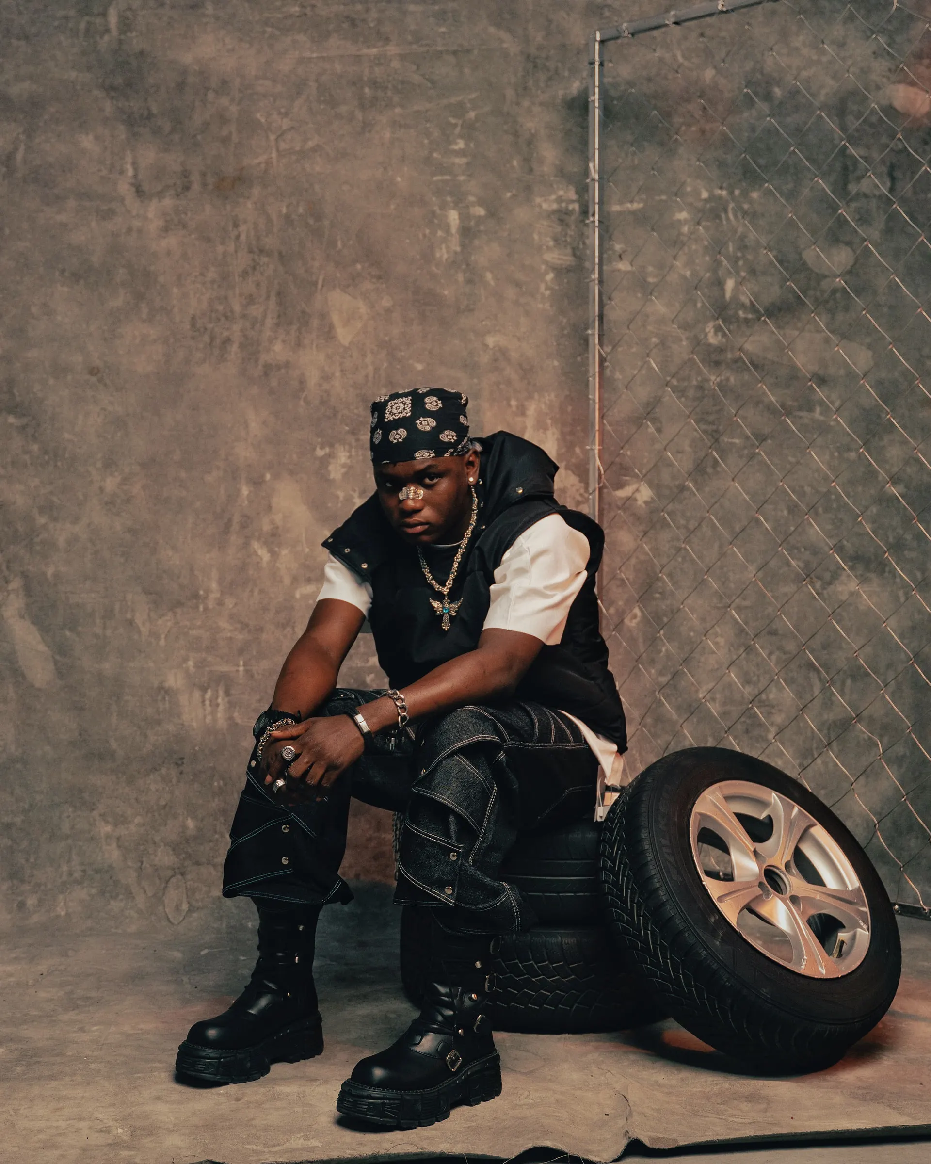 Ayo Maff sits on stacked tyres beside a chain-link fence, wearing a black padded vest over a white tee and looking toward the camera.