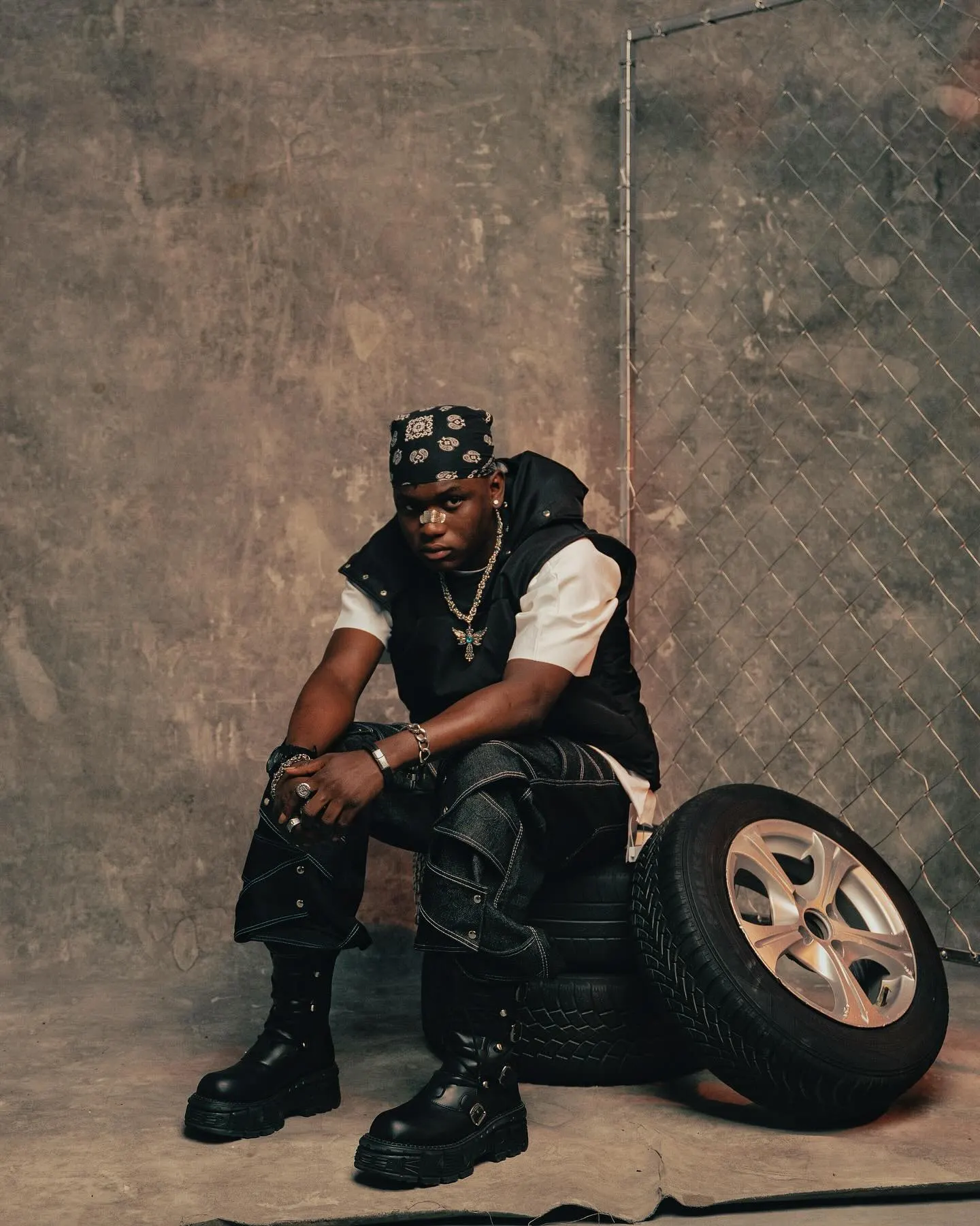 Musician in a black puffer vest over a white tee, seated on stacked tires beside a chain-link gate, elbows on knees against a mottled concrete wall.