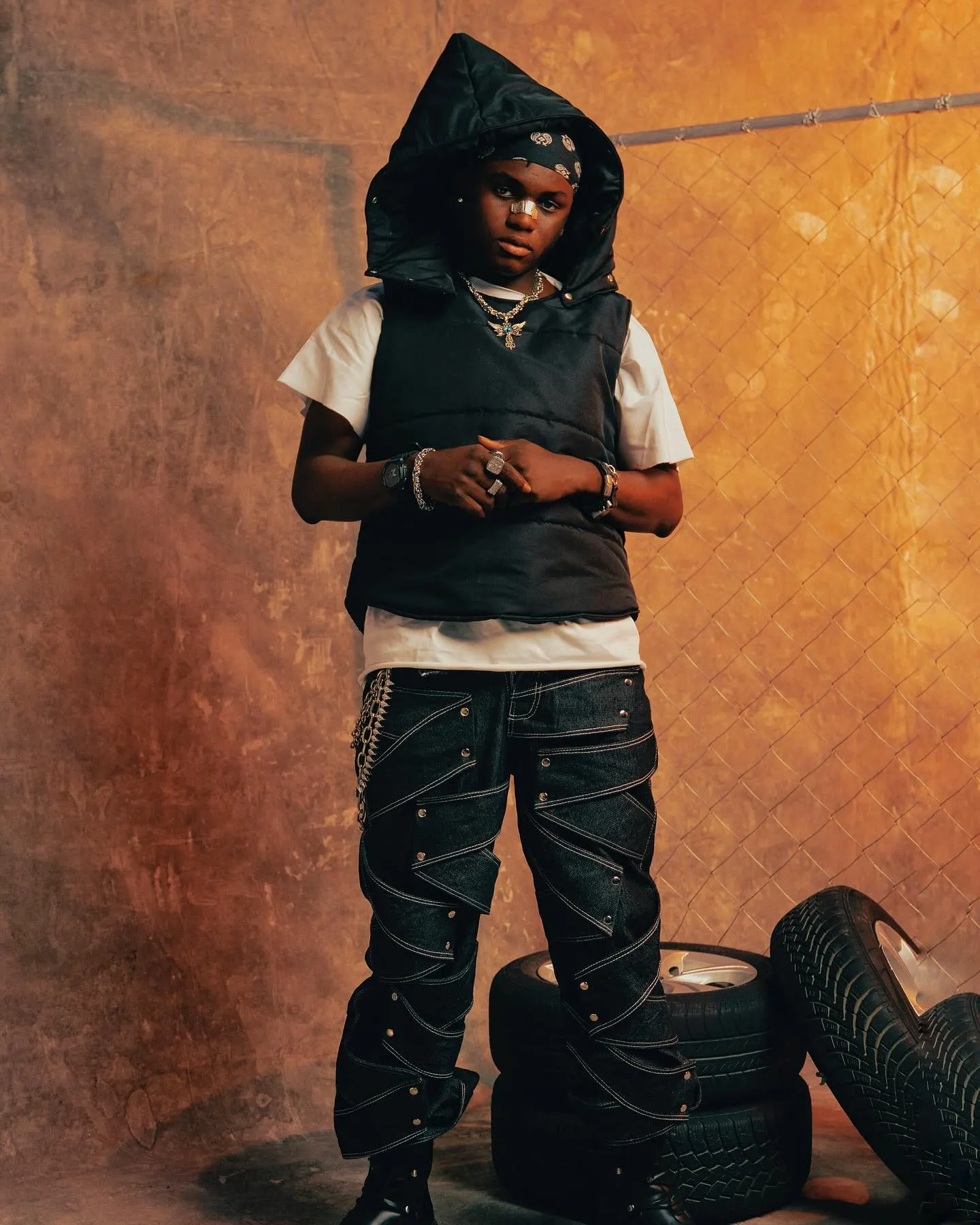 Ayo Maff in a hooded black puffer vest over a white tee, paisley bandana peeking beneath the hood, standing on stacked tyres against a rust-toned wall and chain-link fence.