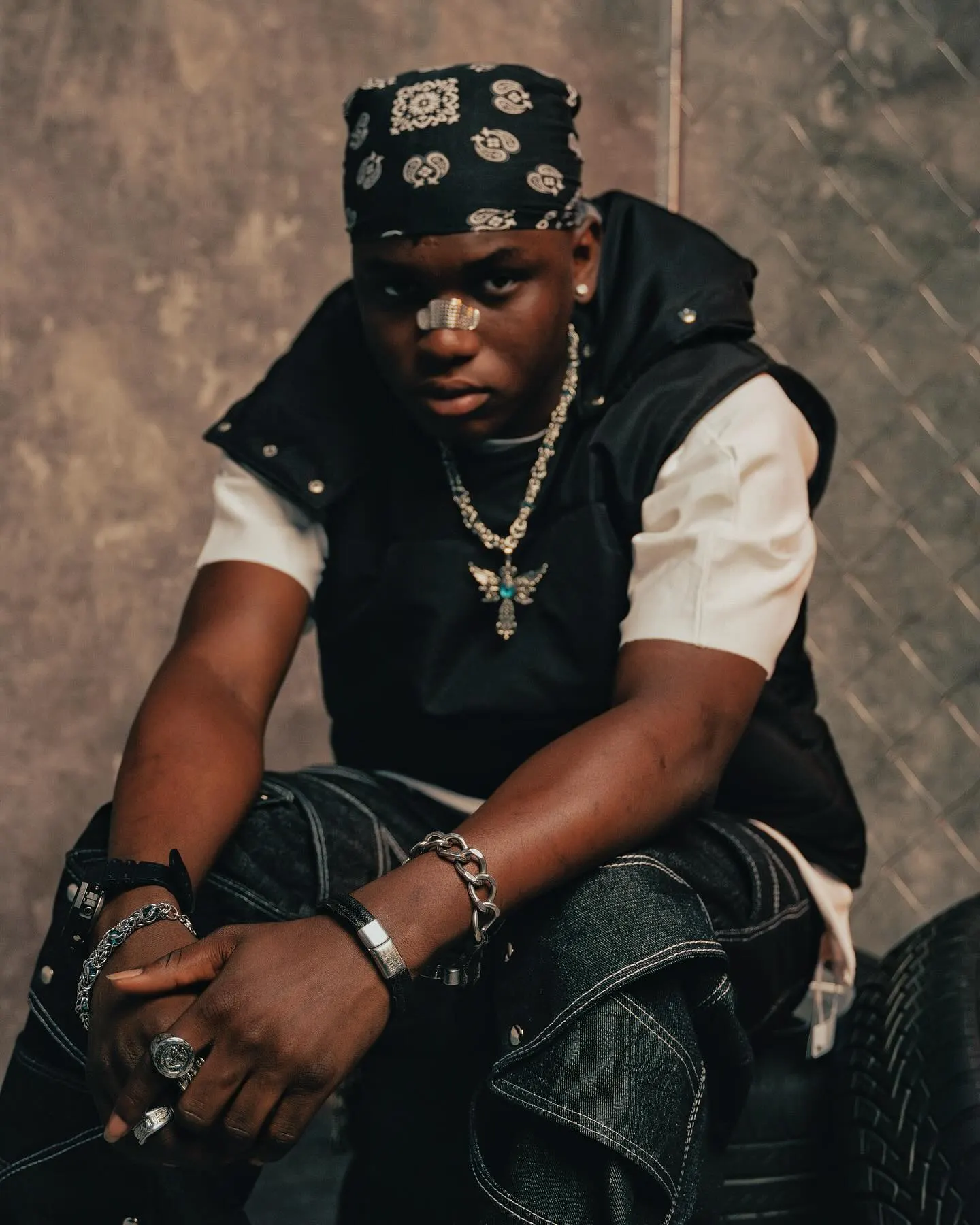 Ayo Maff crouched forward with arms crossed over his knees, paisley bandana and plaster across his nose, silver chains and chunky bracelets catching the light against chain-link fence.
