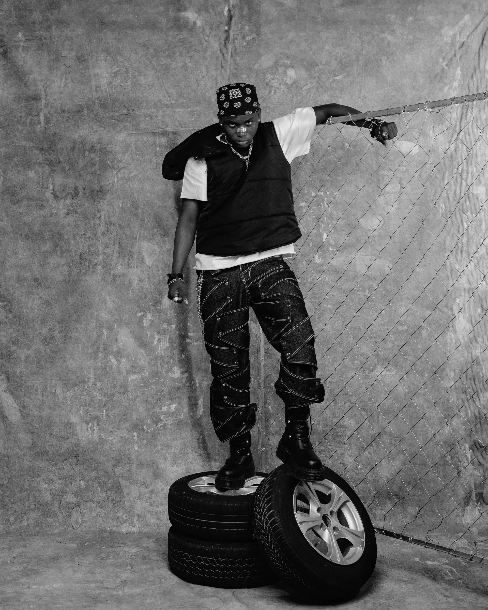 Black-and-white photograph of Ayo Maff standing on stacked tyres, one forearm resting along a chain-link fence.
