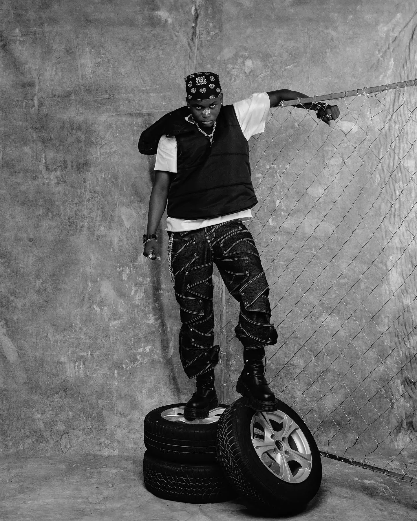 Black-and-white frame of Ayo Maff balanced atop a stack of tyres, one arm gripping the chain-link fence, in bandana, black vest and studded criss-cross jeans.