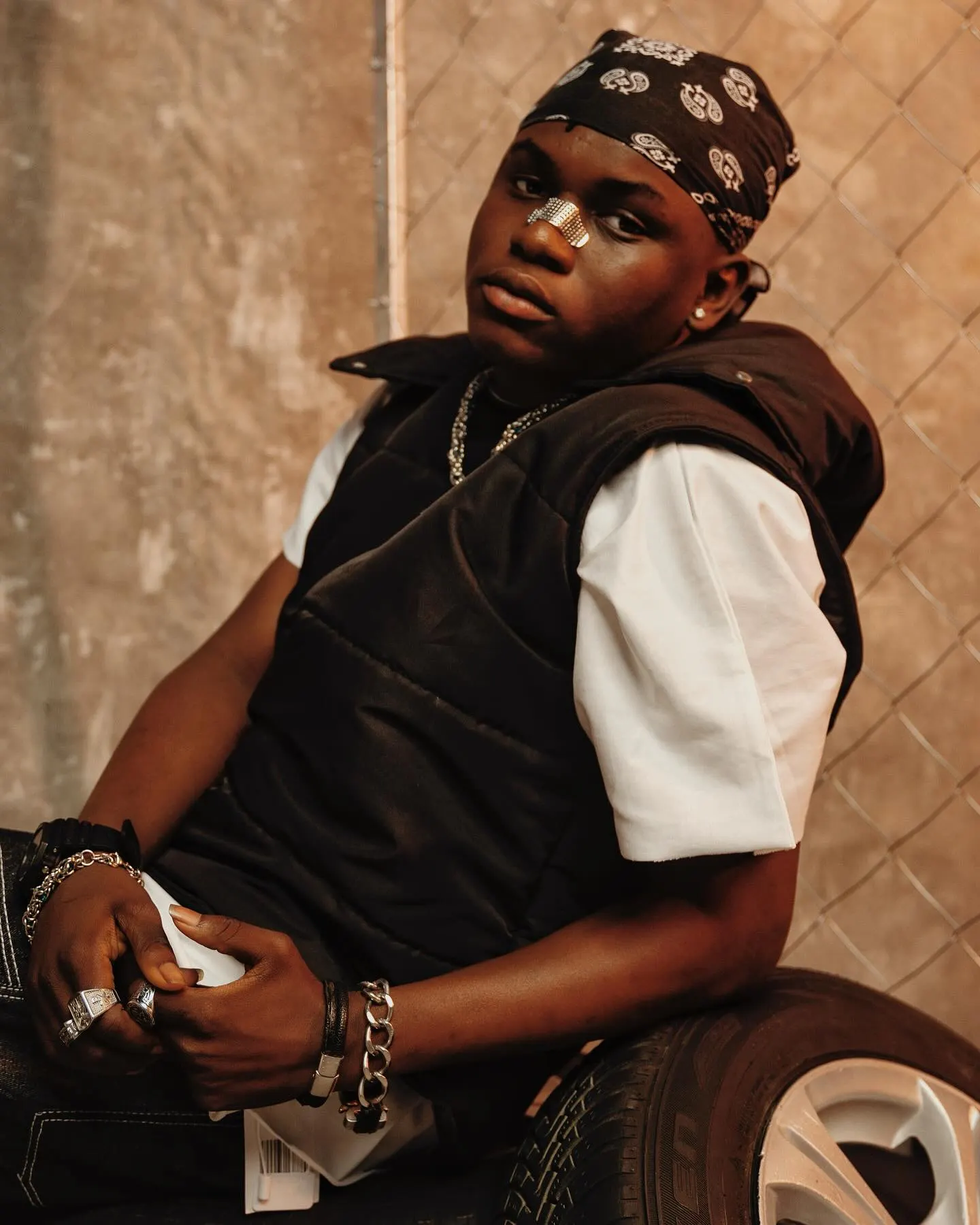 Ayo Maff seated on a tyre in a black paisley bandana and quilted vest, plaster on his nose, gazing off past the camera against a warm plaster wall and fence.