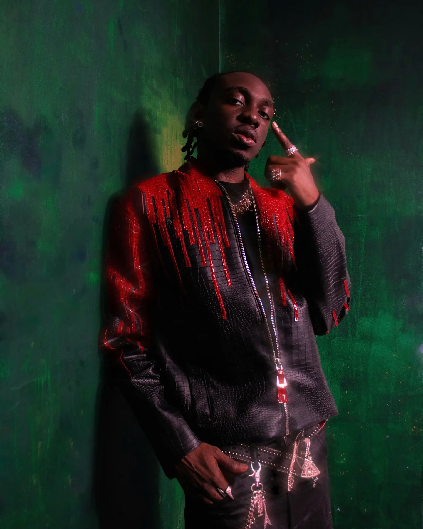 Producer in a black snakeskin jacket dripping with red beaded fringe, one ringed finger raised to his temple against a mottled green wall.