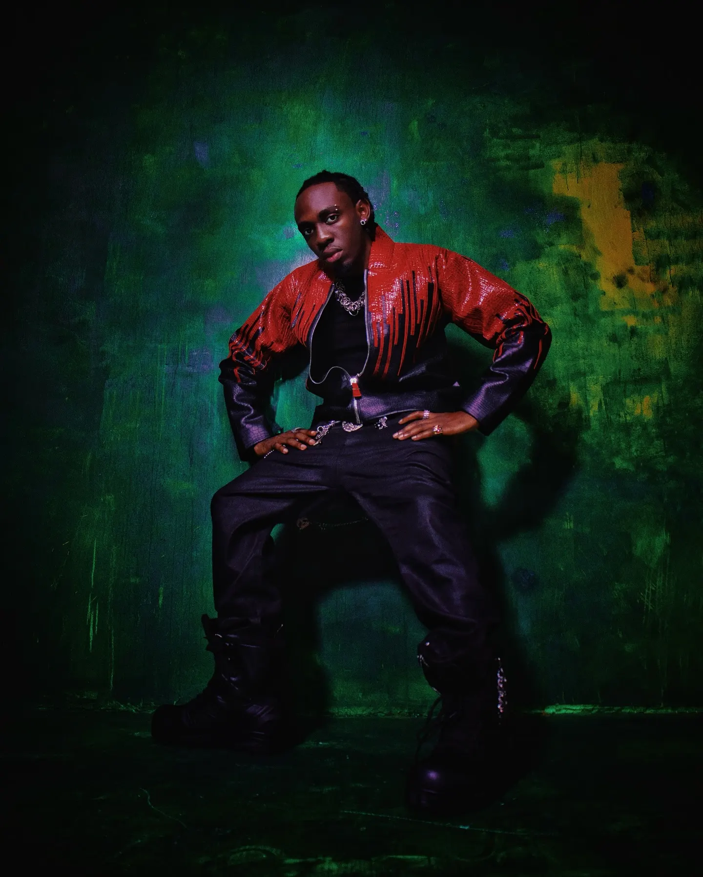 Seated figure in a red-fringed leather jacket and heavy black boots, hands planted on his knees before a paint-streaked green backdrop.