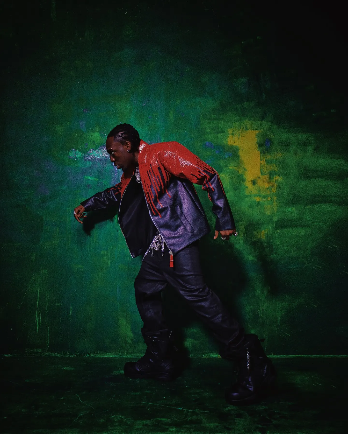 Full-length profile mid-stride in a red-and-black leather jacket, boots dragging across a green-washed studio floor.