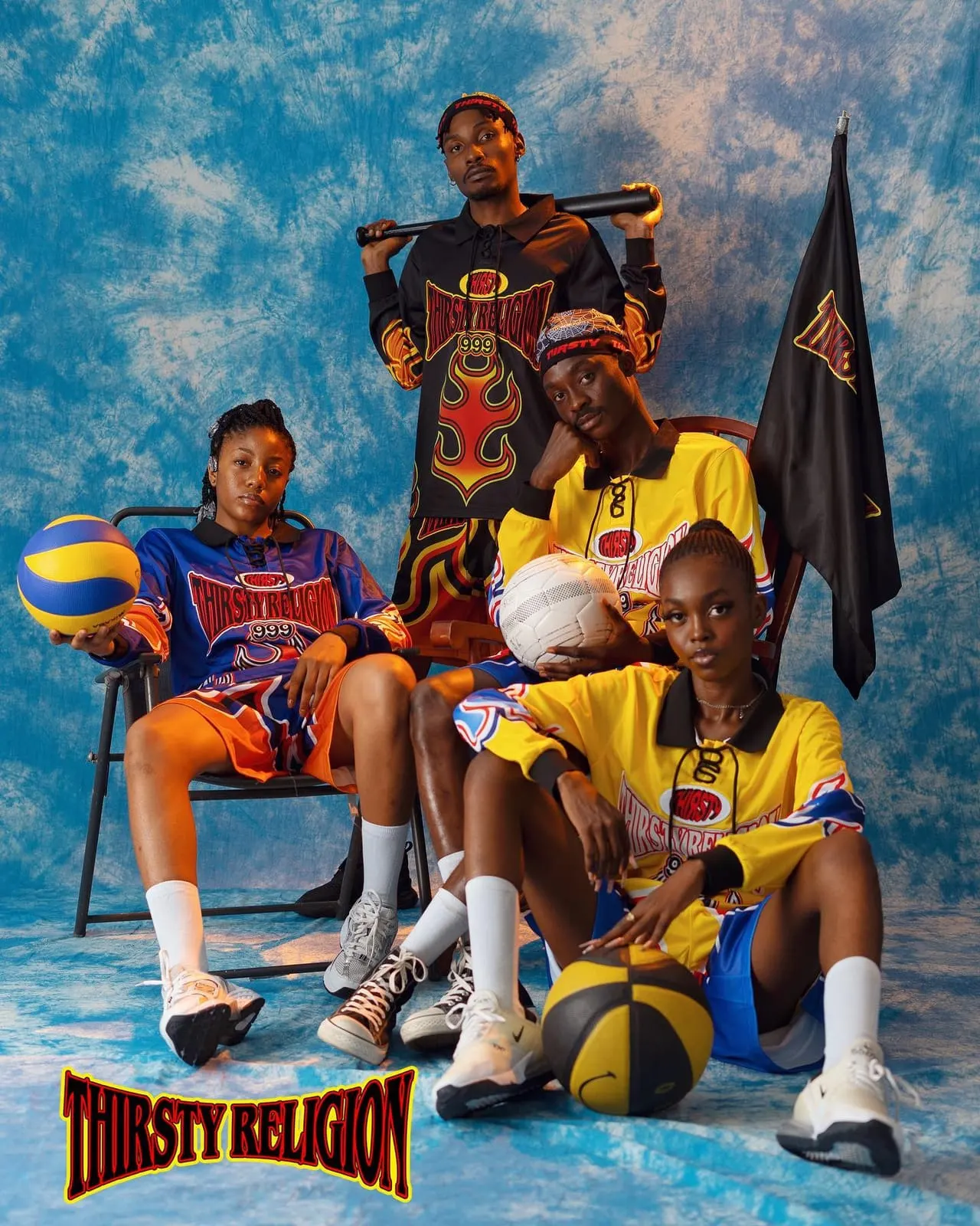 Four models in flame-print Thirsty Religion jerseys pose with basketballs and a team flag against blue tie-dye.