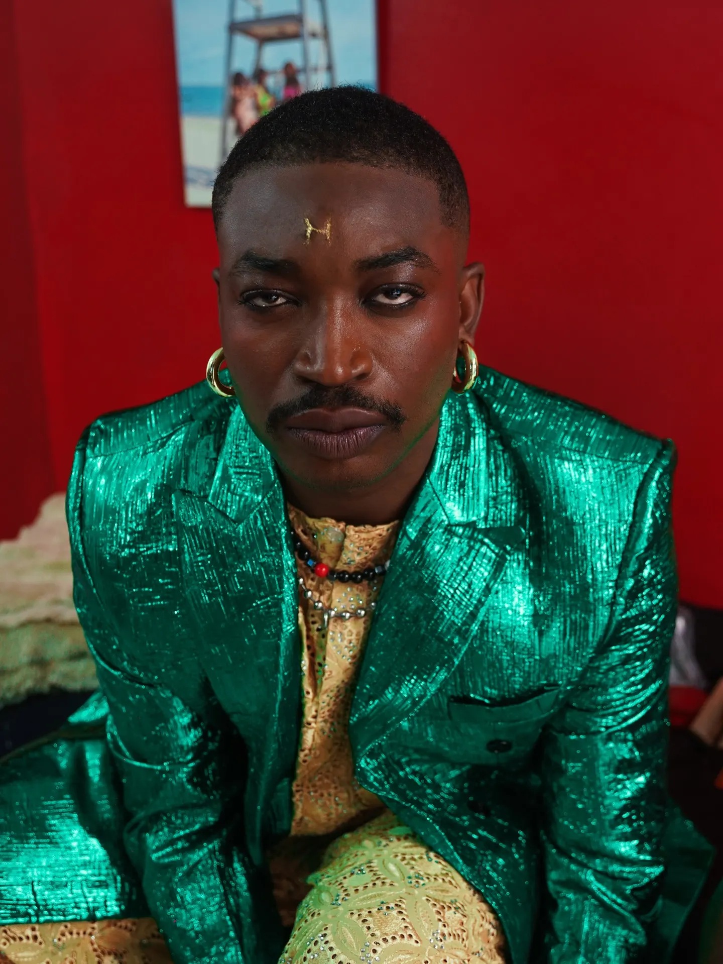 Musician in a metallic emerald-green blazer over a gold lace shirt, staring straight into the lens from a low angle against a deep red wall.