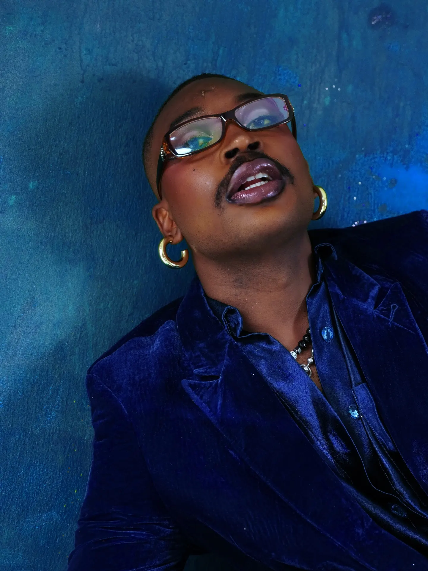 Musician in a blue velvet blazer and tinted glasses tips his head back against a mottled blue wall, gold hoop earrings catching the light.