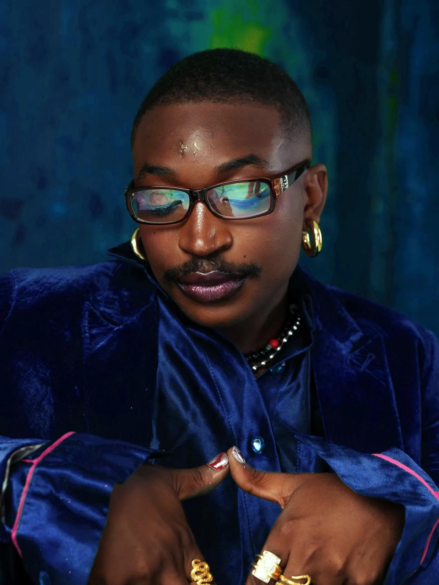 Musician in a blue velvet suit with pink piping leans forward over crossed arms, gold rings and beaded necklace against a blue-green painted backdrop.
