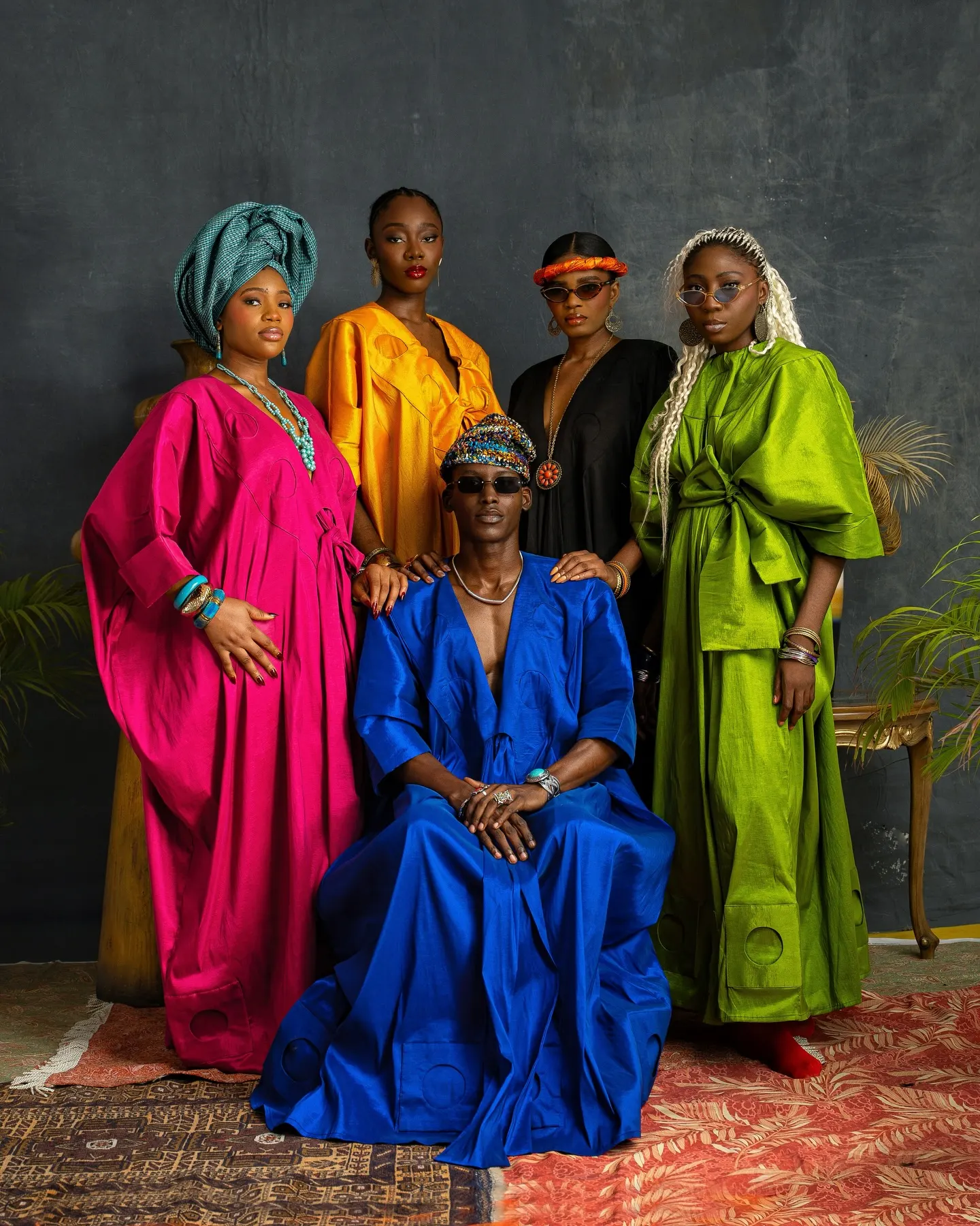 Five models in jewel-toned silk kaftans — magenta, marigold, black, green and cobalt — pose in a tight group against a mottled grey studio canvas.