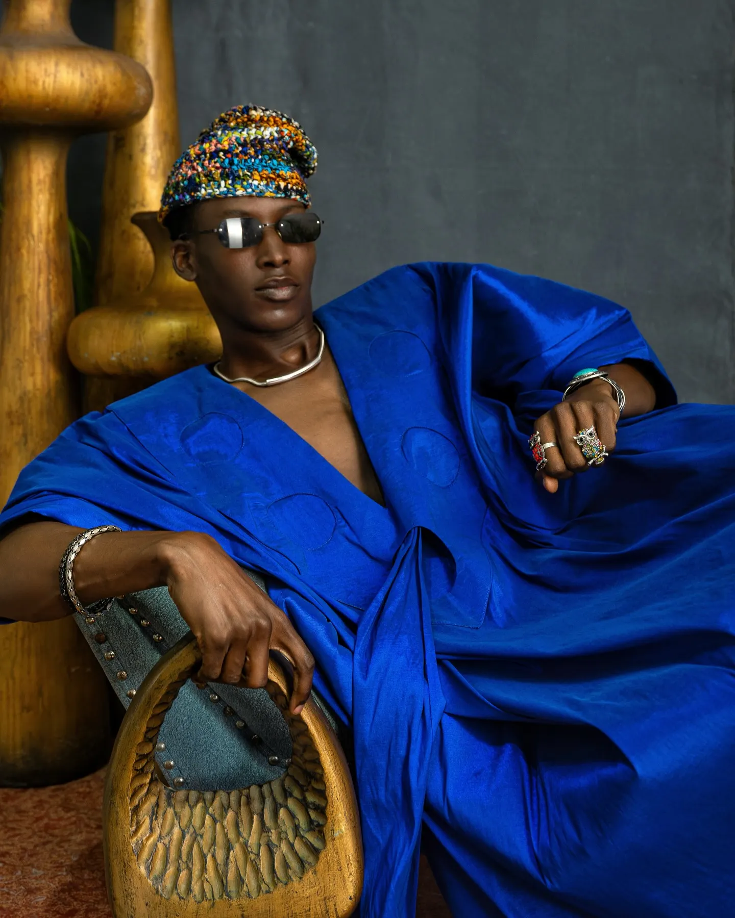 Model in a voluminous cobalt-blue kaftan, beaded cap and mirrored sunglasses reclines with one arm draped over a carved wooden chair.