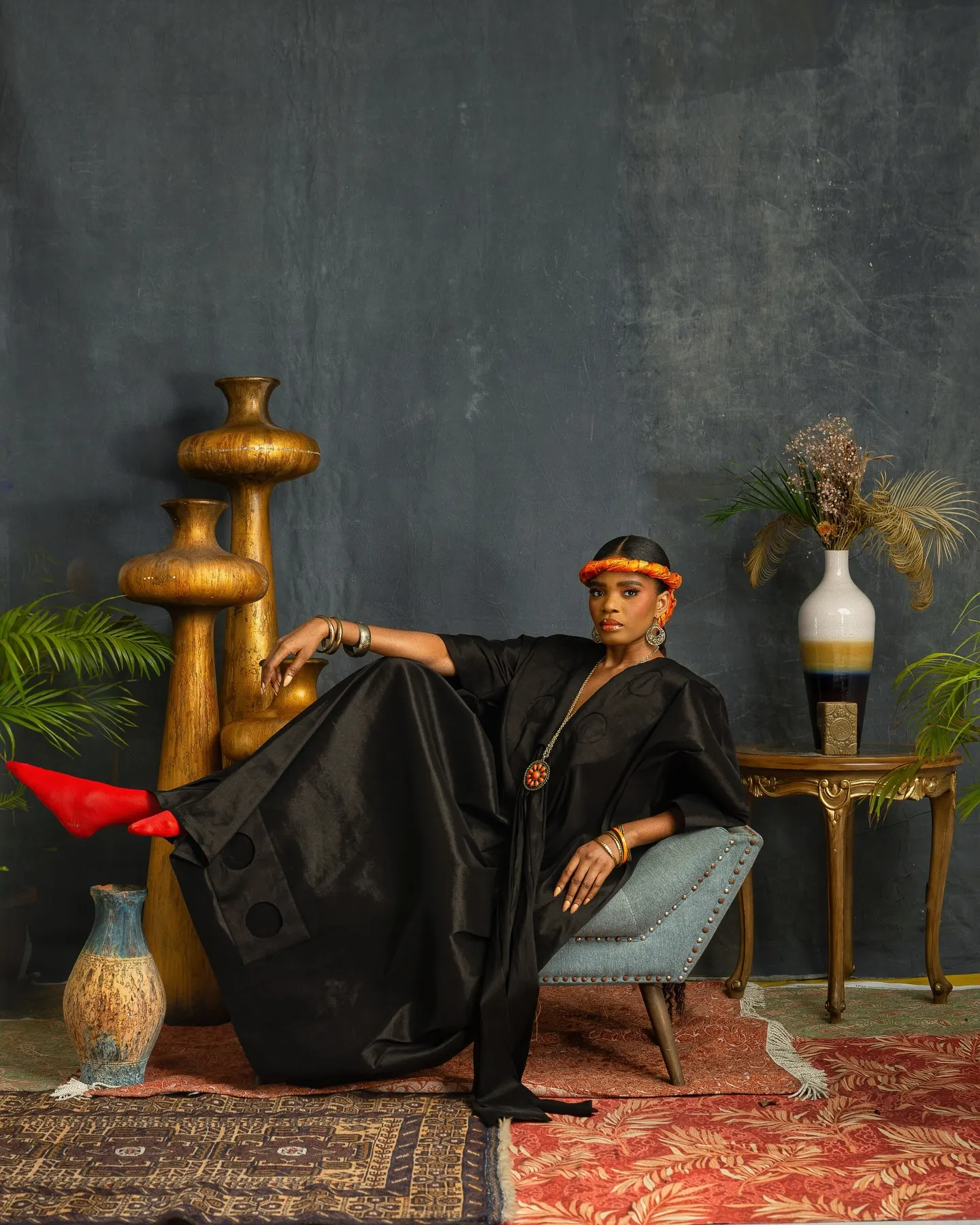 Model in a cobalt-blue kaftan with a bow-tied neckline crosses her wrists at her collarbone, a braided orange-and-yellow hairpiece falling past her shoulder.