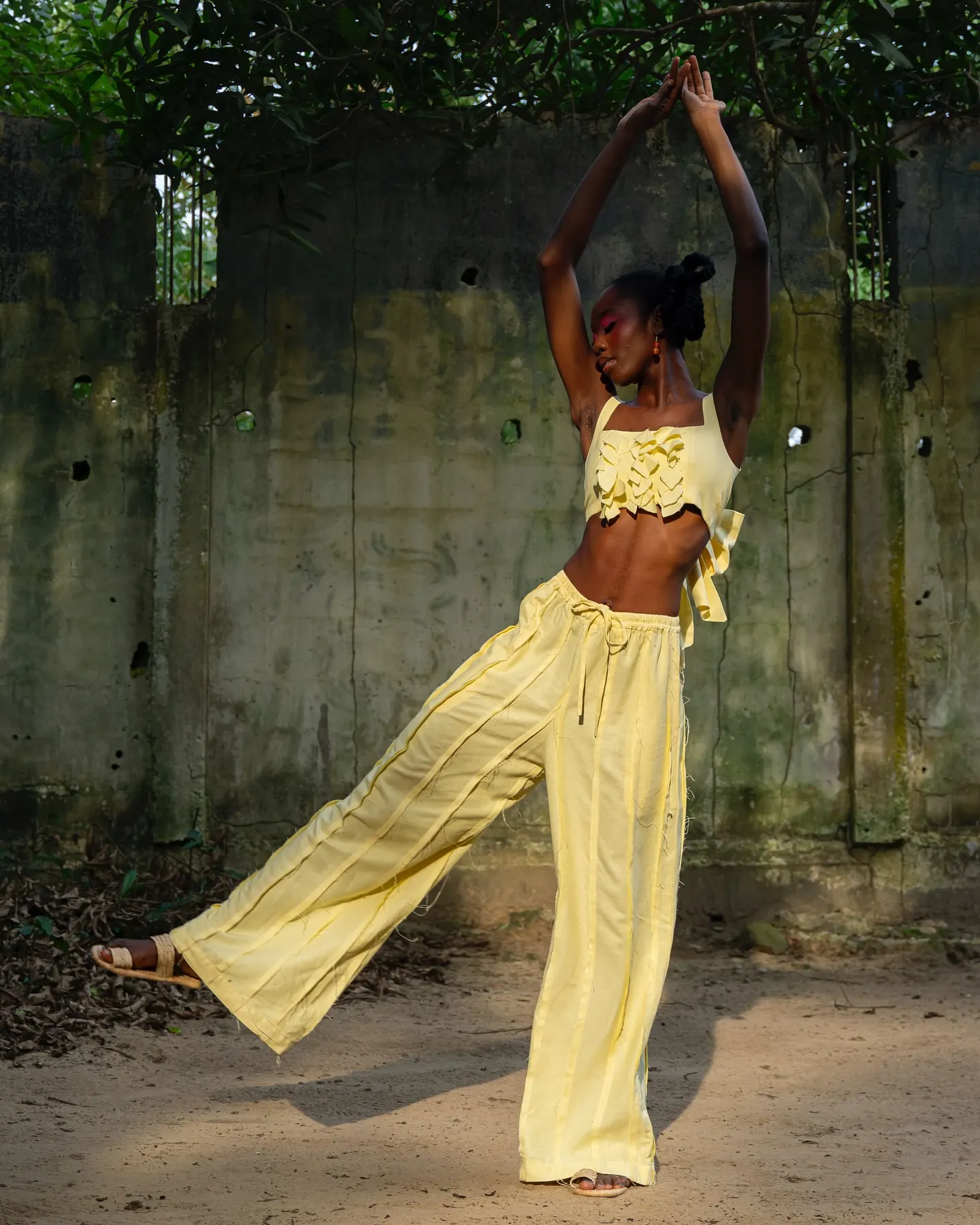 Model in a pale-yellow ruffled crop top and wide-leg trousers arches mid-dance with arms raised and one leg kicked out before a mossy concrete wall.