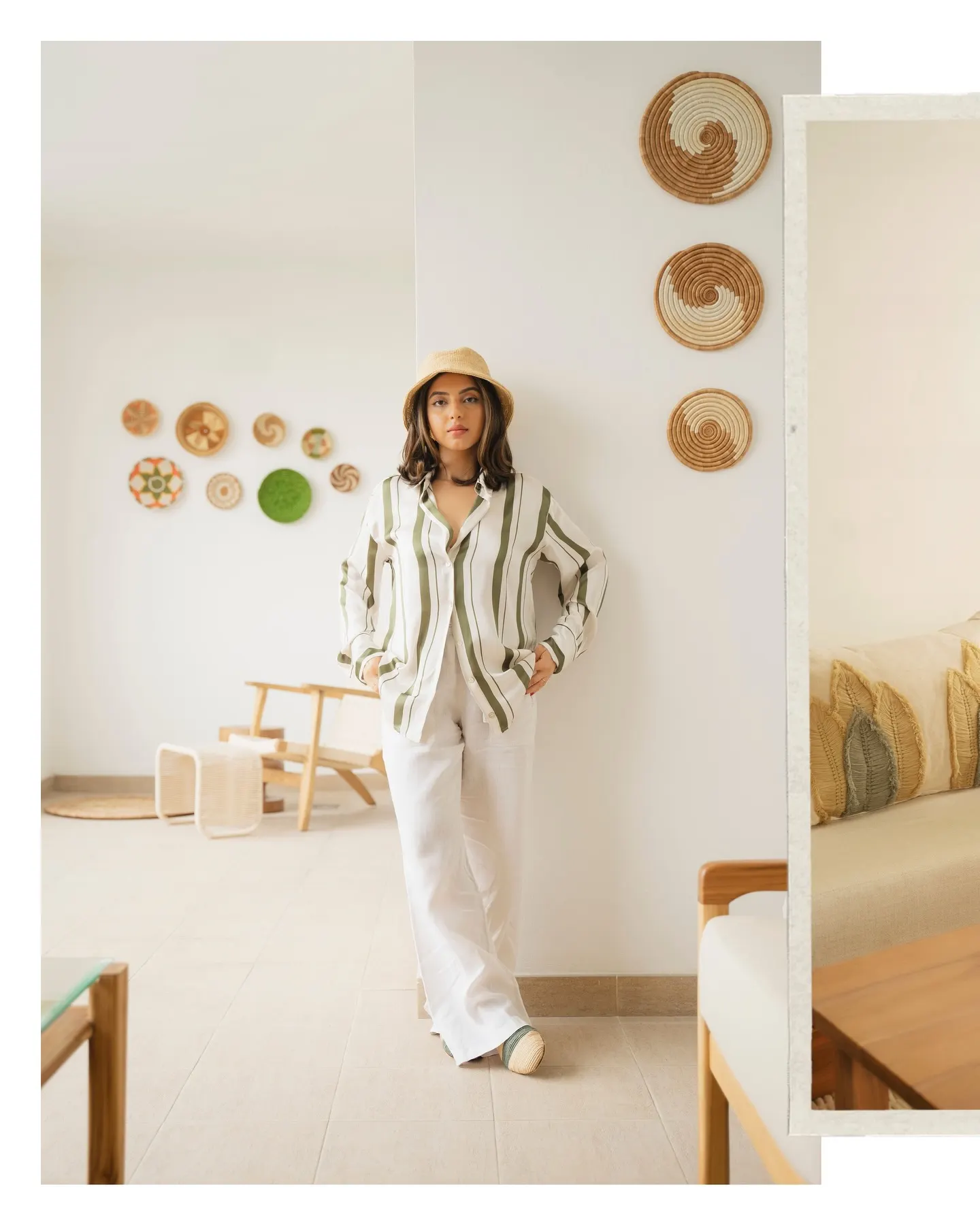 Woman in an olive-striped shirt, white trousers and straw hat stands full-length in a sunlit living room hung with woven wall baskets.